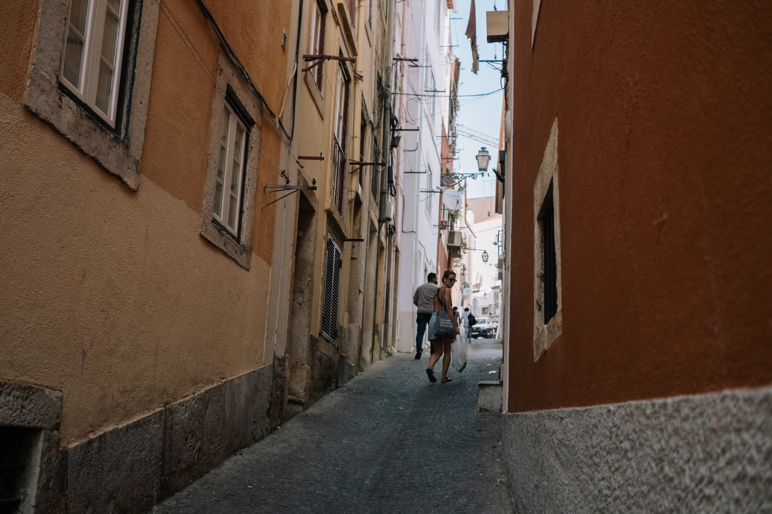People walking in a narrow alleyway between colorful buildings with small windows, satellite dishes, and street lamps.