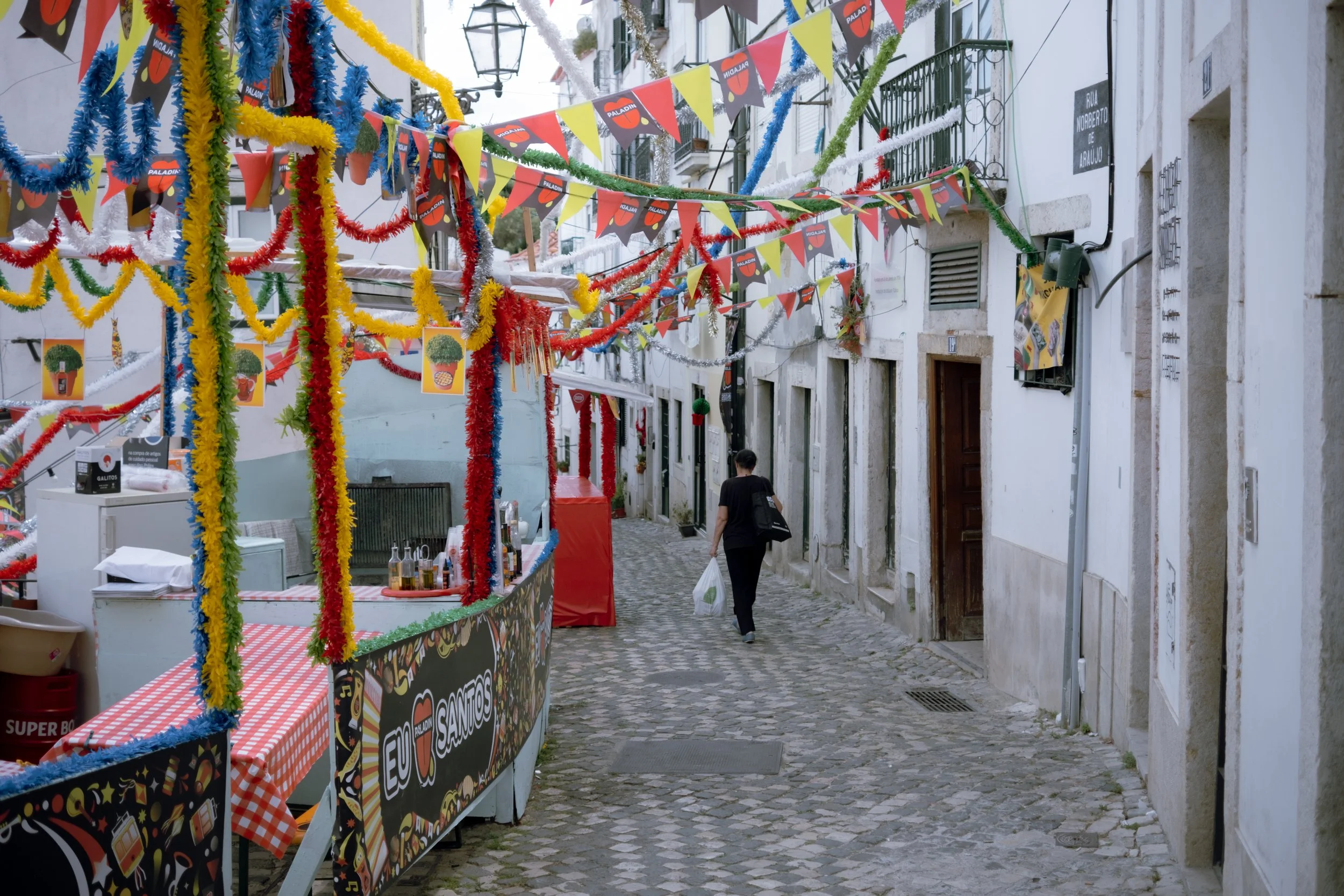 A narrow cobblestone street decorated with colorful banners and streamers for a festive celebration. A person walks down the street carrying shopping bags, with small vendor stalls on the side.