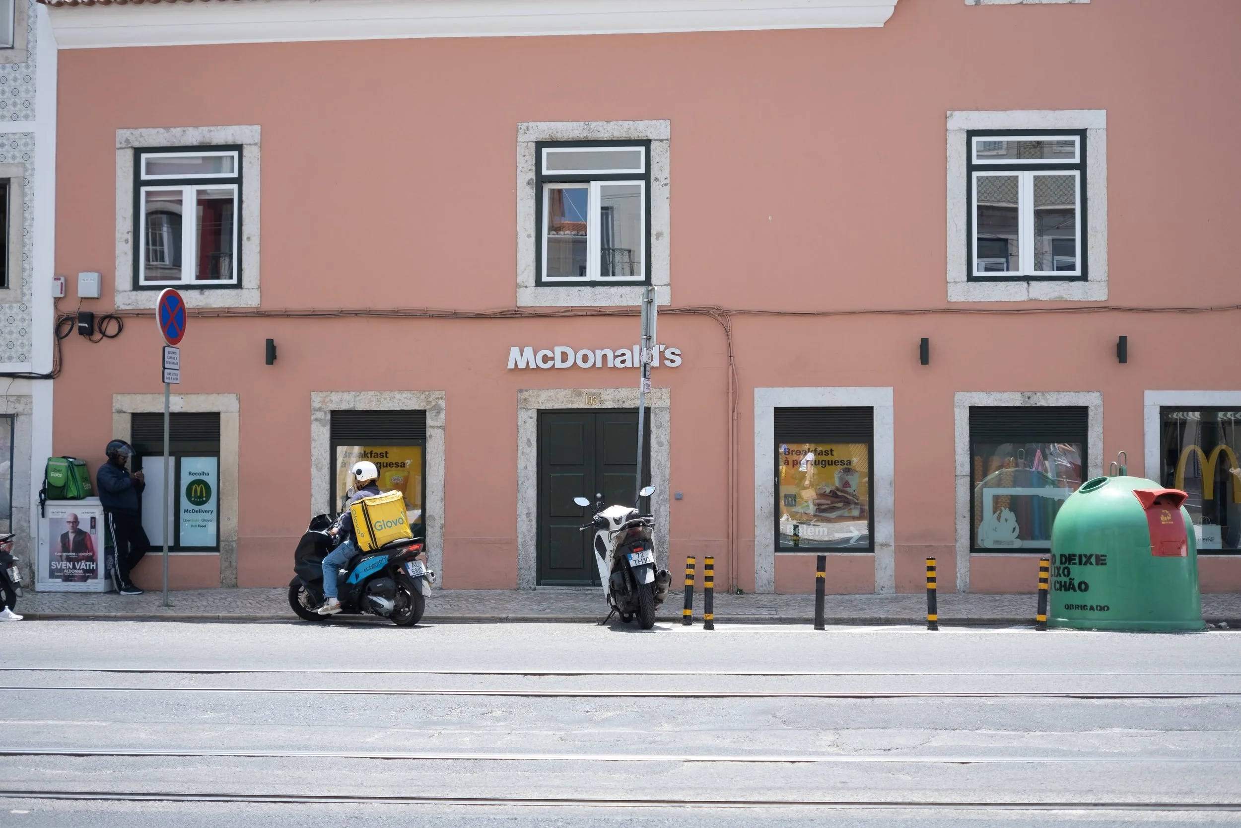 Street view of a pink building with McDonald's sign, two motorcycle riders, a pedestrian, a McDonald's delivery person, a green and red recycling bin, and tram tracks in the foreground.