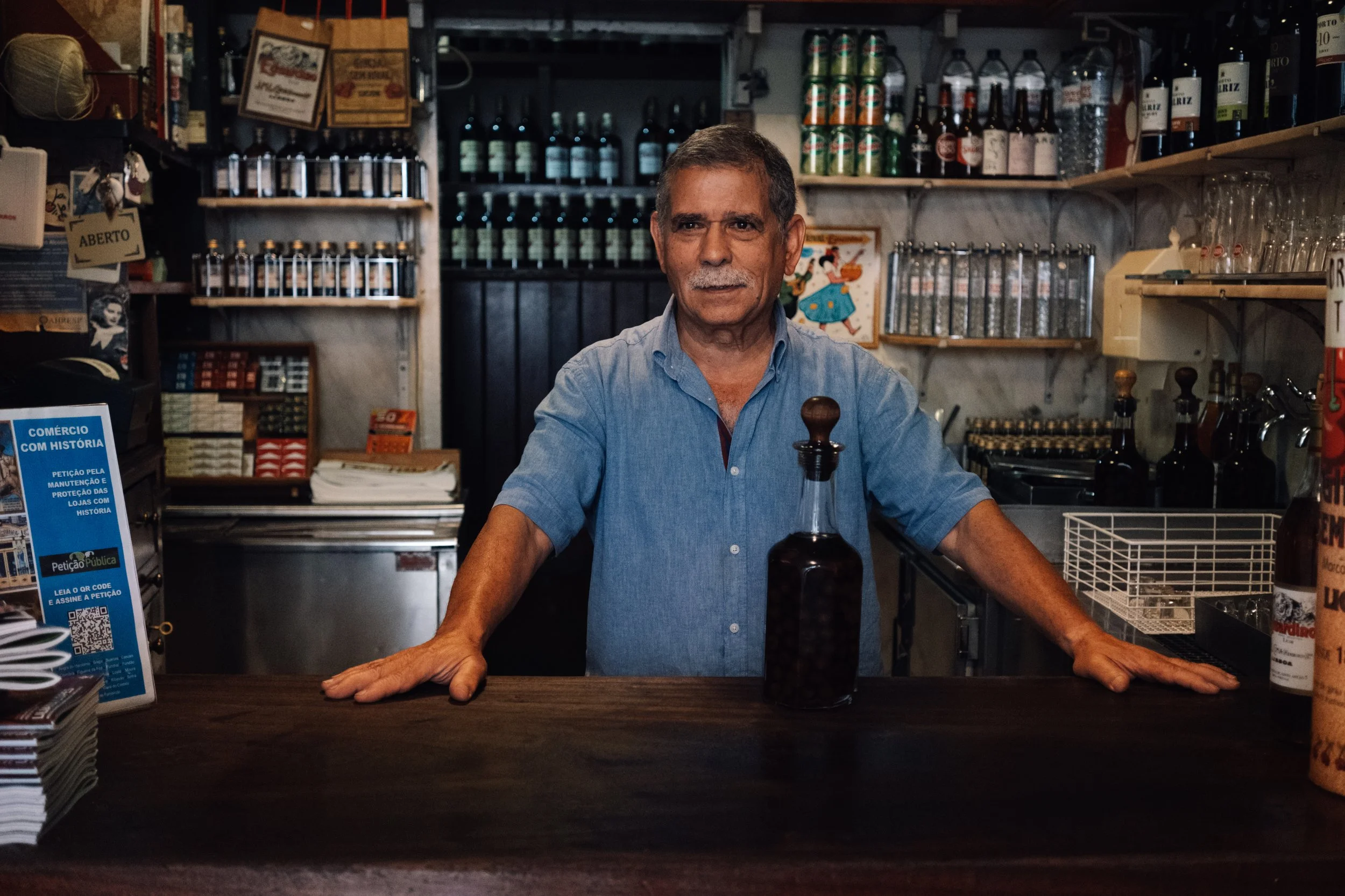 An older man standing behind a bar counter with various bottles and cans of beverages in the background.