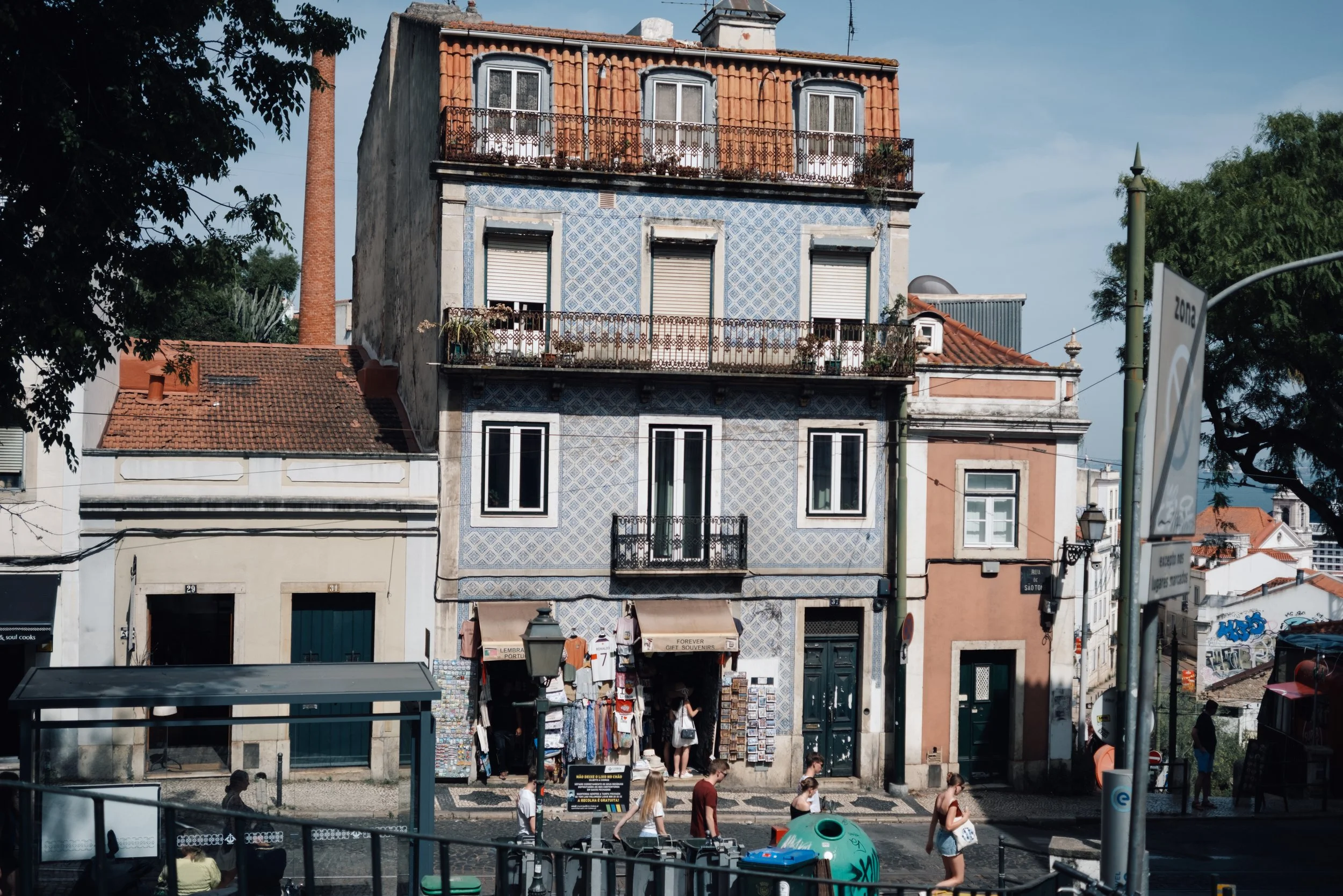 Street scene with a tall, old building with a blue patterned facade and balconies, surrounded by other buildings, trees, and people walking on the sidewalk.