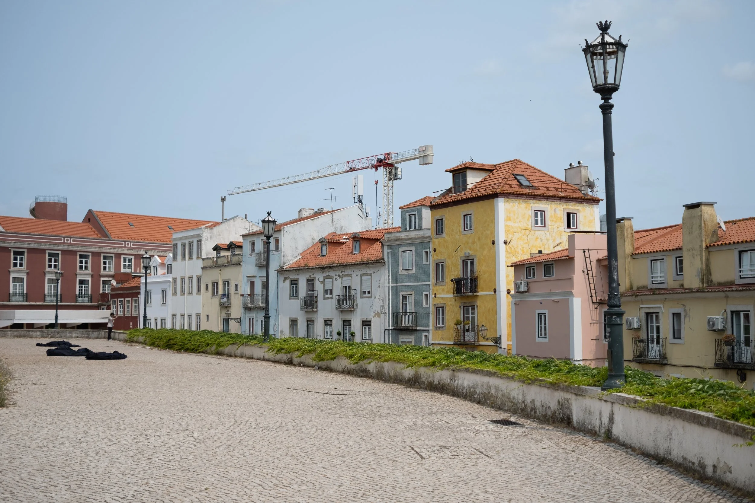 Colorful European-style buildings with red tile roofs along a cobblestone walkway, black street lamps, and a construction crane in the background.