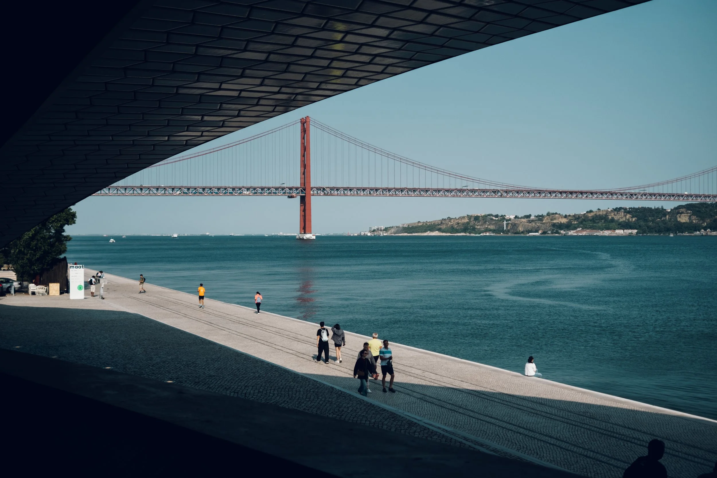 View of a large red suspension bridge over blue water, with people walking and sitting along the waterfront promenade, under a shaded walkway.