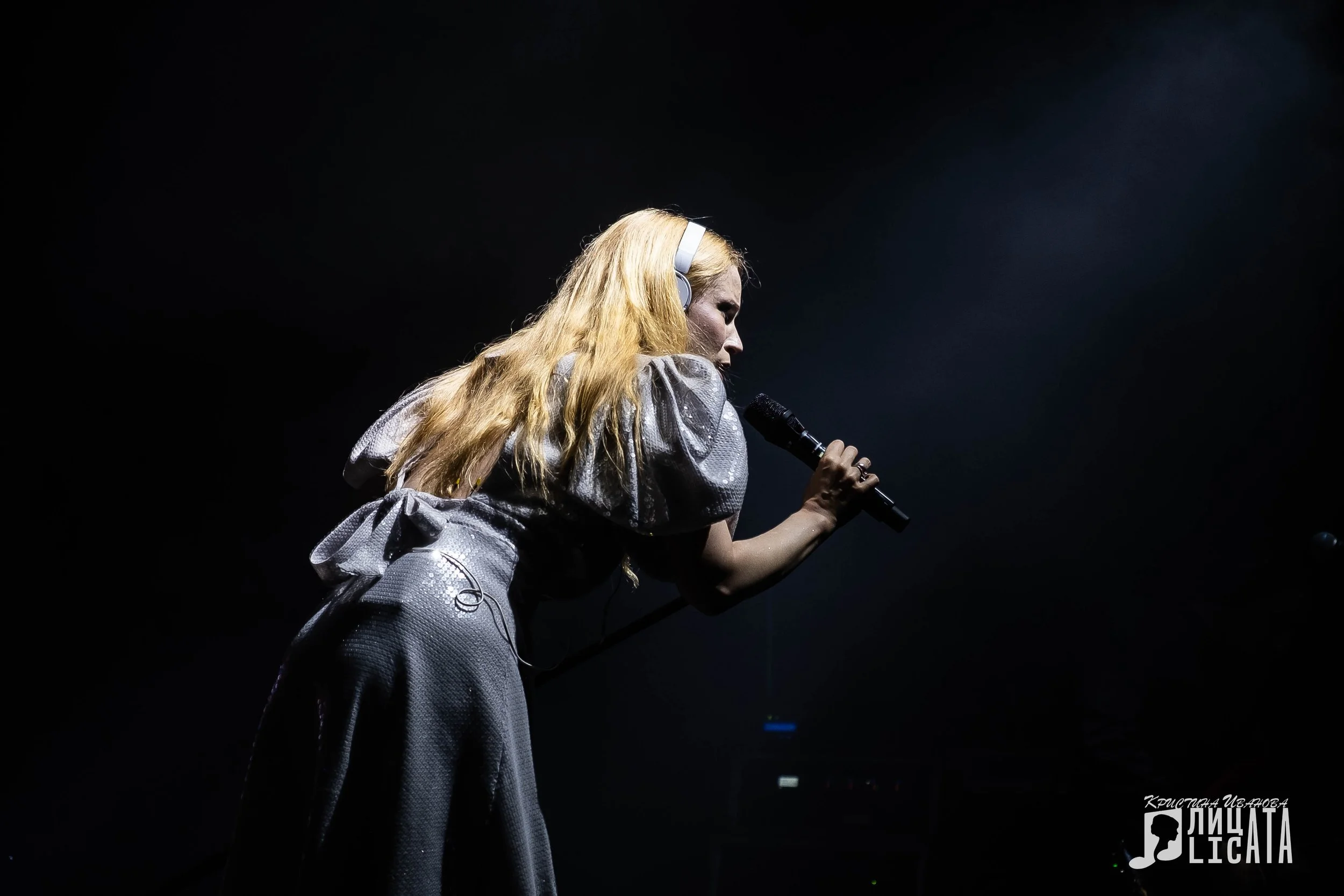 A female performer with long blonde hair wearing a shiny silver outfit and headphones, singing into a microphone on a dark stage, with a DJ or band member partially visible in the background.