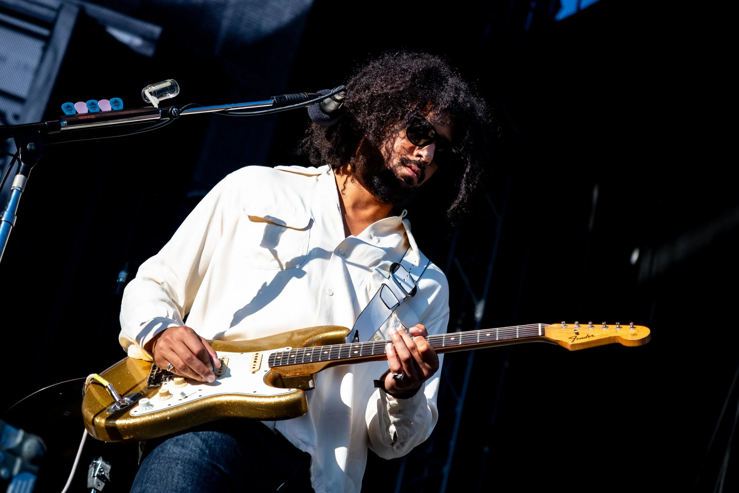 Musician playing an electric guitar on stage, wearing sunglasses and a white shirt.