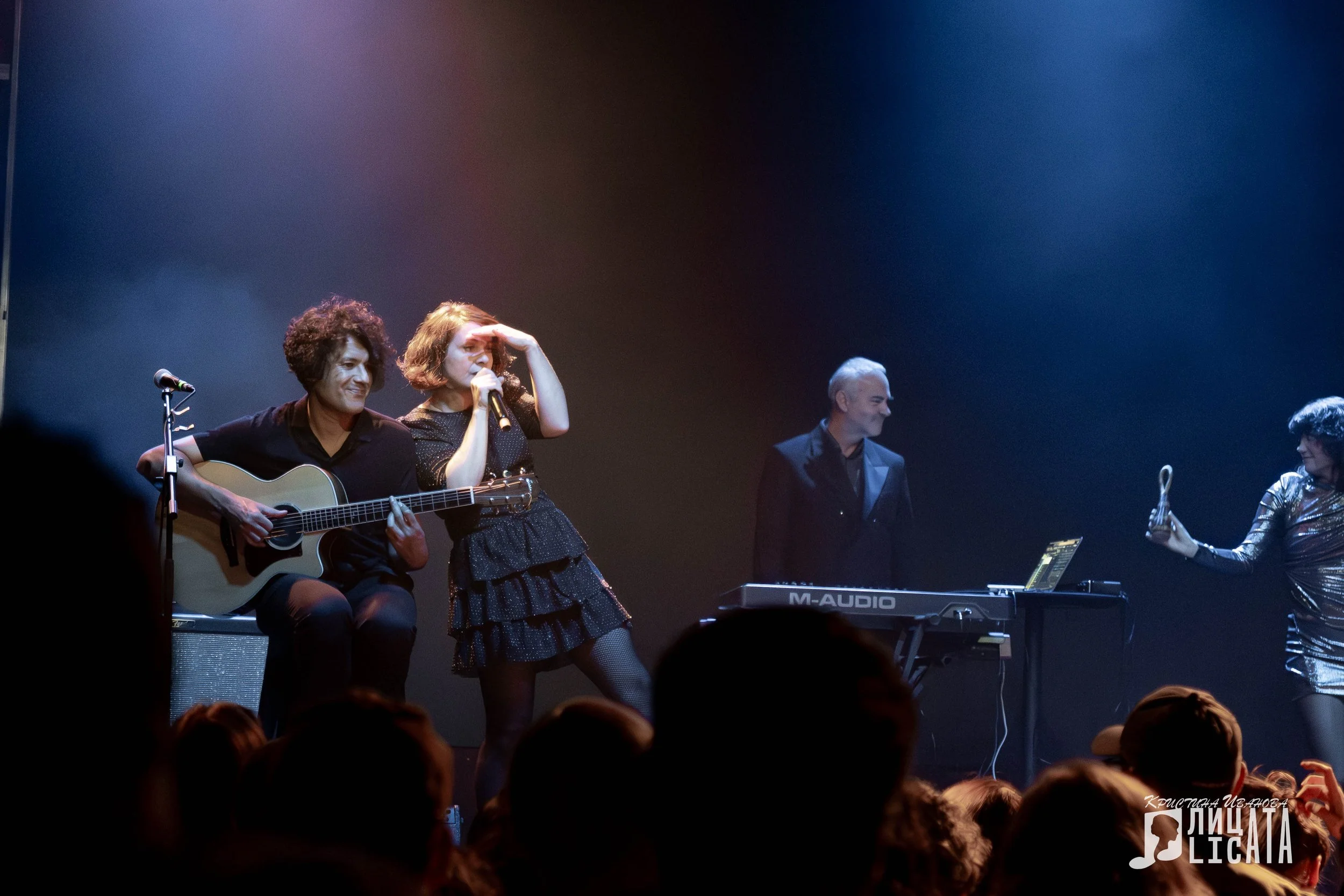 A live concert with a female singer performing on stage, accompanied by musicians, under colorful stage lights, with an audience watching and taking photos.
