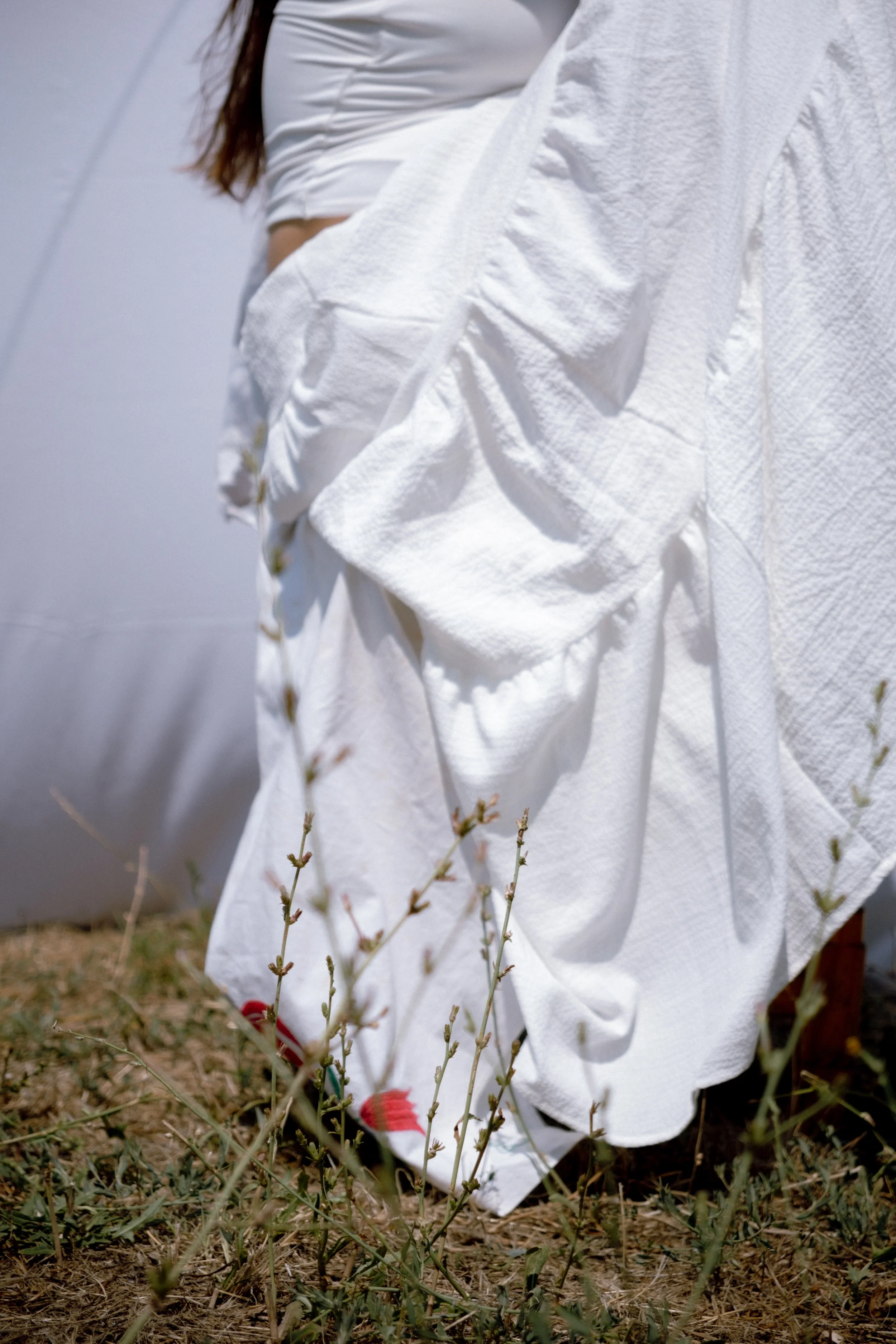 A person wearing a long white dress kneeling on dry grass with small plants, with a light-colored tent or fabric in the background.