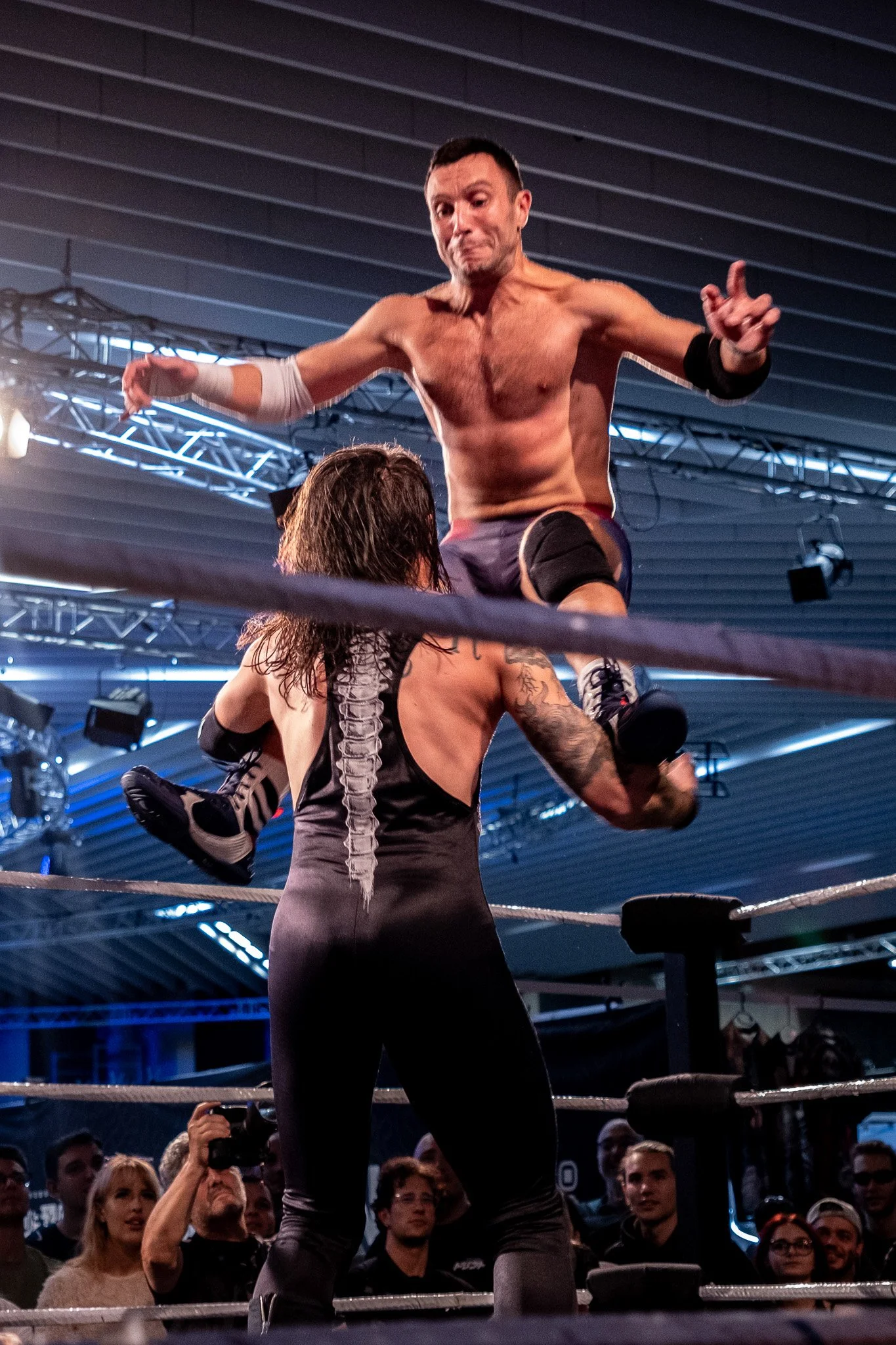 A wrestling match showing one wrestler mid-air, jumping on another wrestler in the ring, with audience members watching.