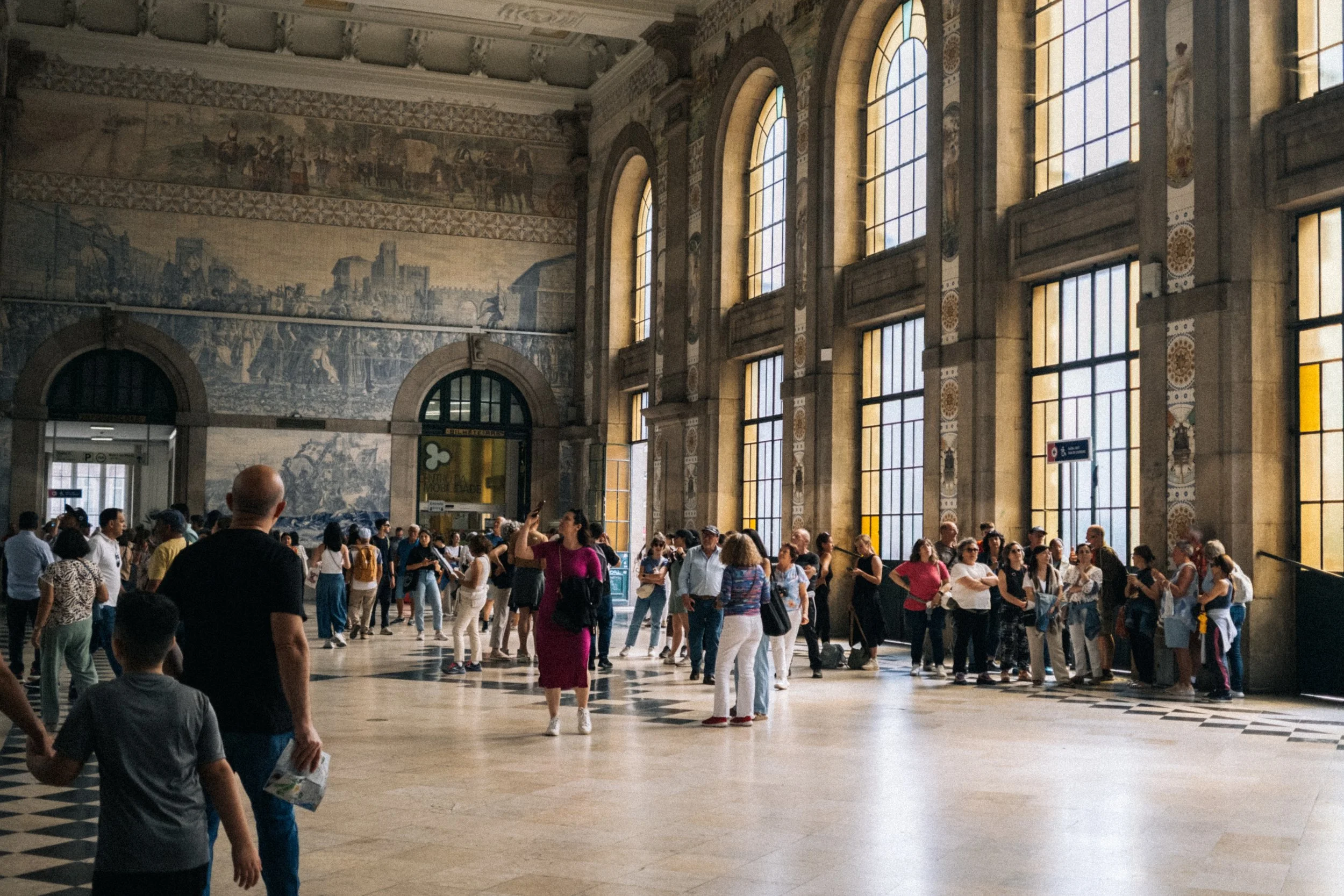 Interior of a grand train station with tall arched windows, murals on the walls, and many people waiting, walking, and talking.