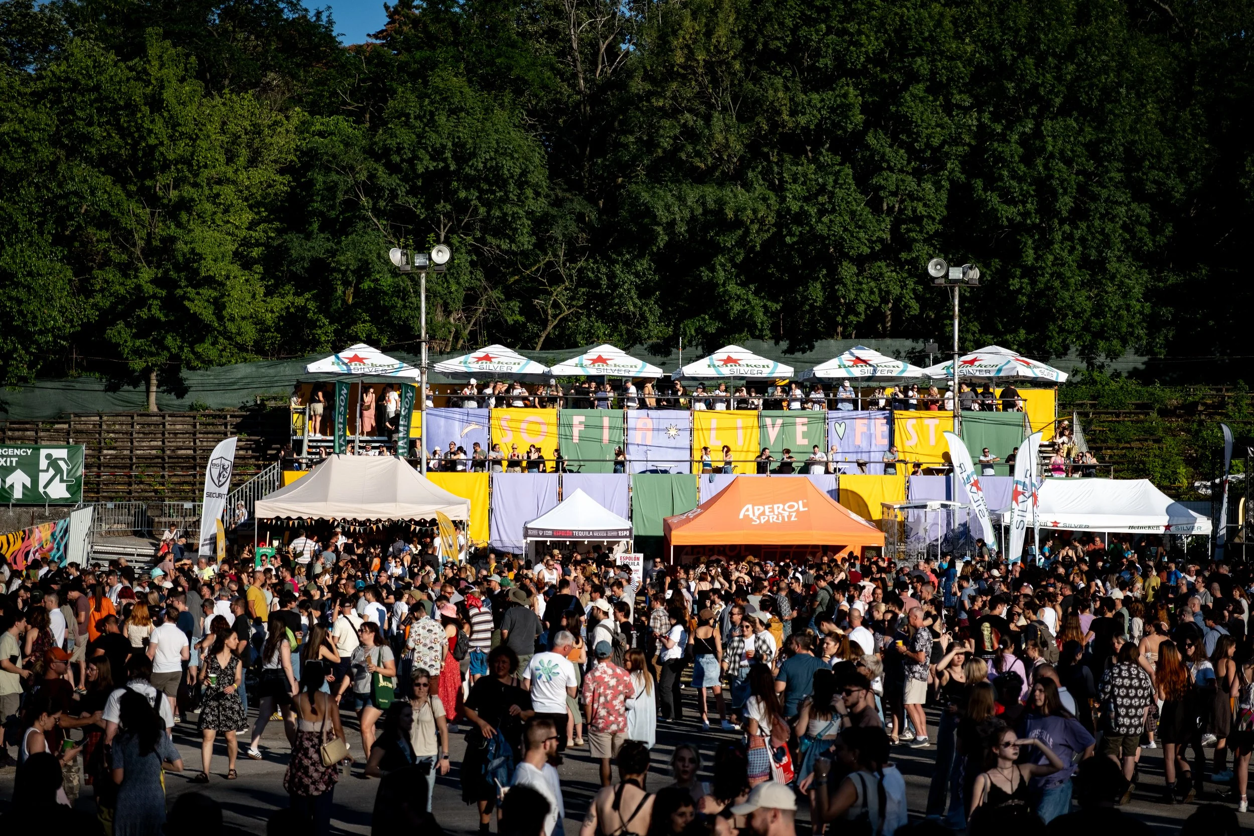 Crowd of people attending an outdoor festival with a stage decorated with colorful banners and umbrellas, surrounded by trees.