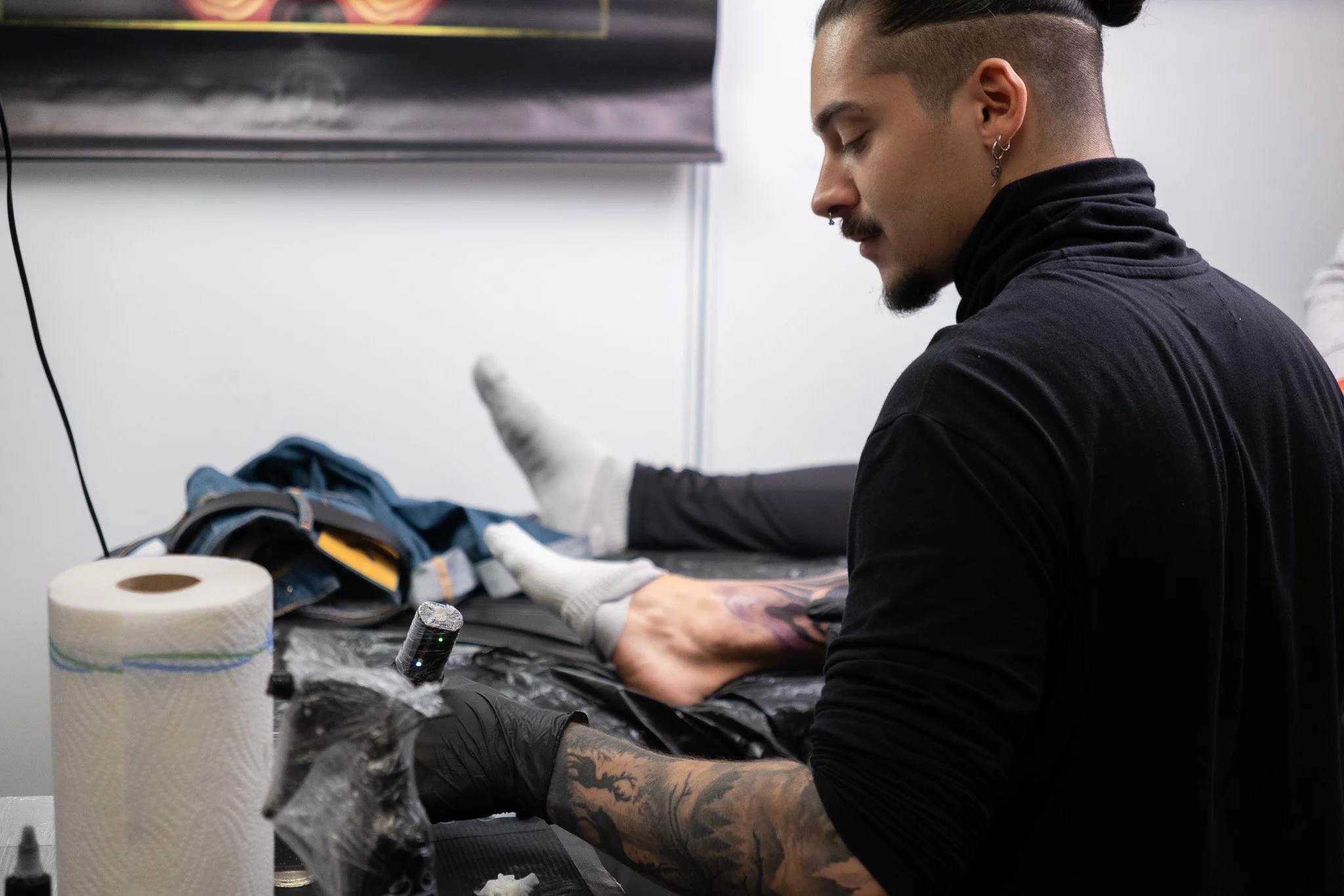Tattoo artist wearing black shirt and gloves tattooing a person's forearm in a tattoo studio, with supplies like paper towels and ink bottles on the table.