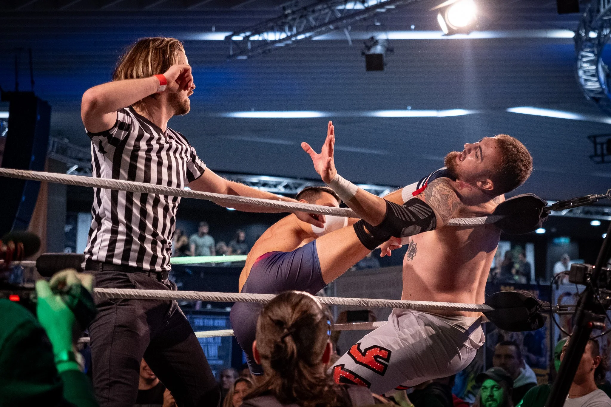 Professional wrestling match where one wrestler delivers a high kick to another, who is caught against the ropes. A referee is present, observing the action, inside a crowded arena.