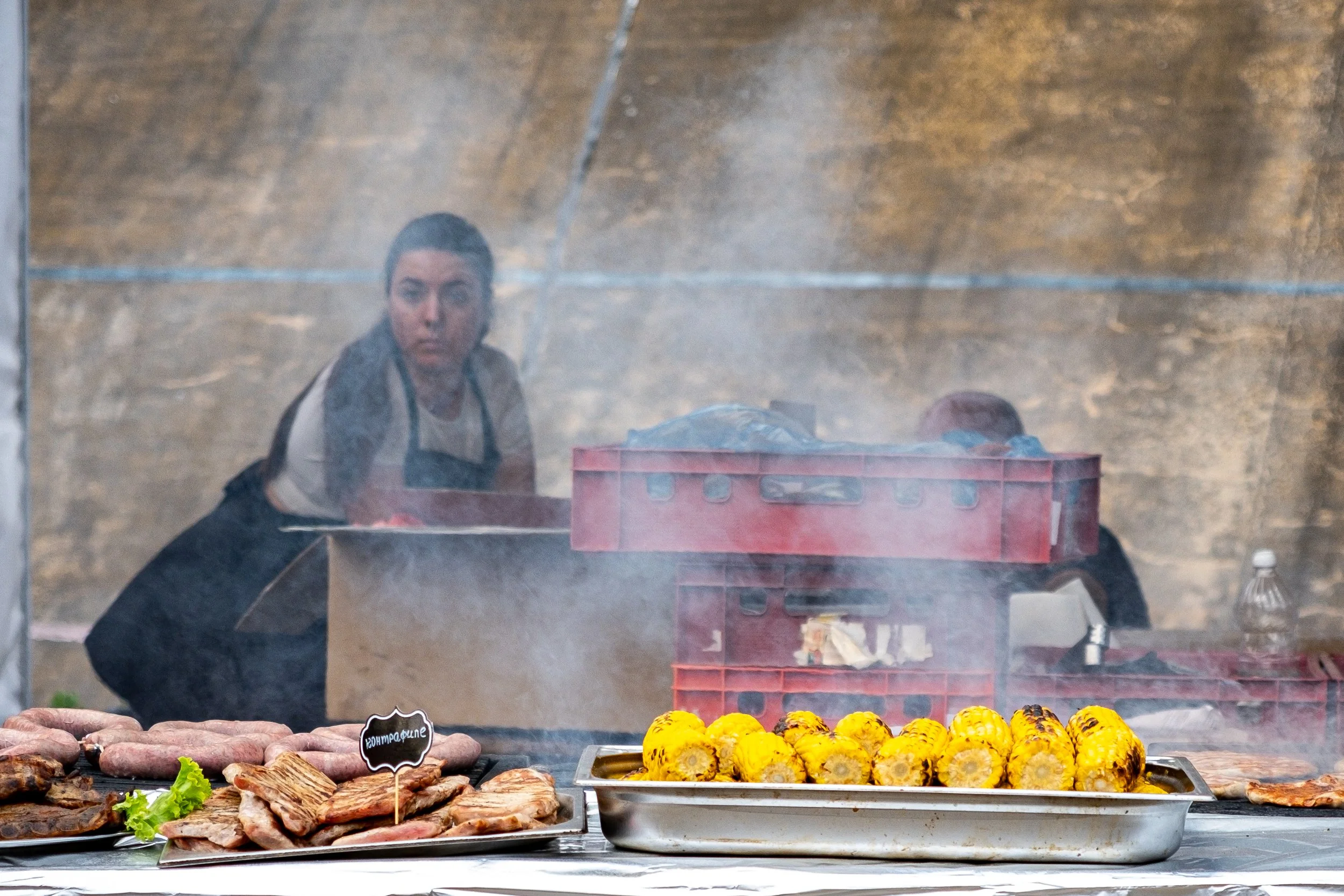A person looking through a grill or smoky cooking area with grilled corn and various meats on trays in the foreground.