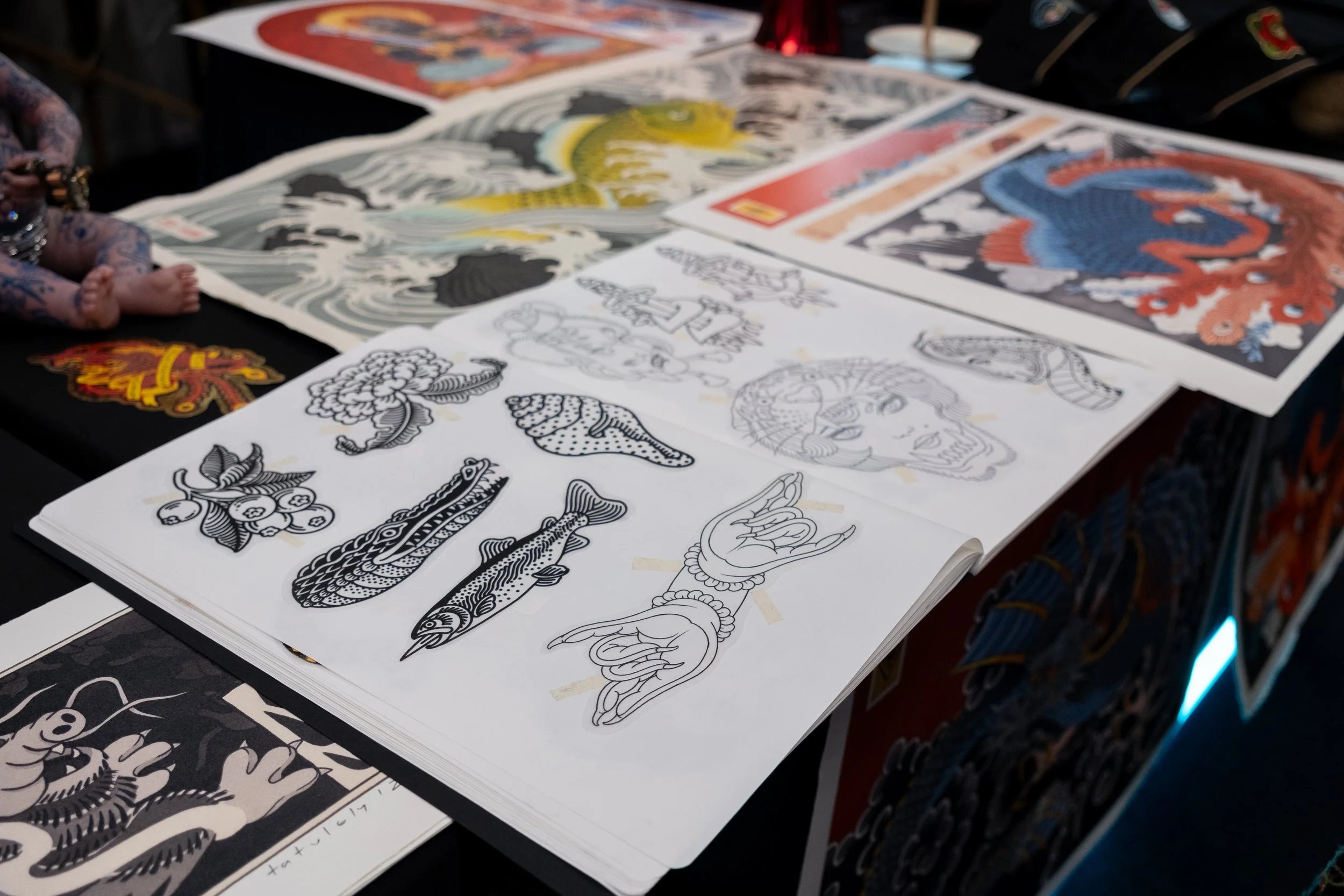 Sheet of tattoo designs featuring traditional style tattoos, including a skull, hand gesture, fish, and flower, displayed on a table at an art or tattoo convention.