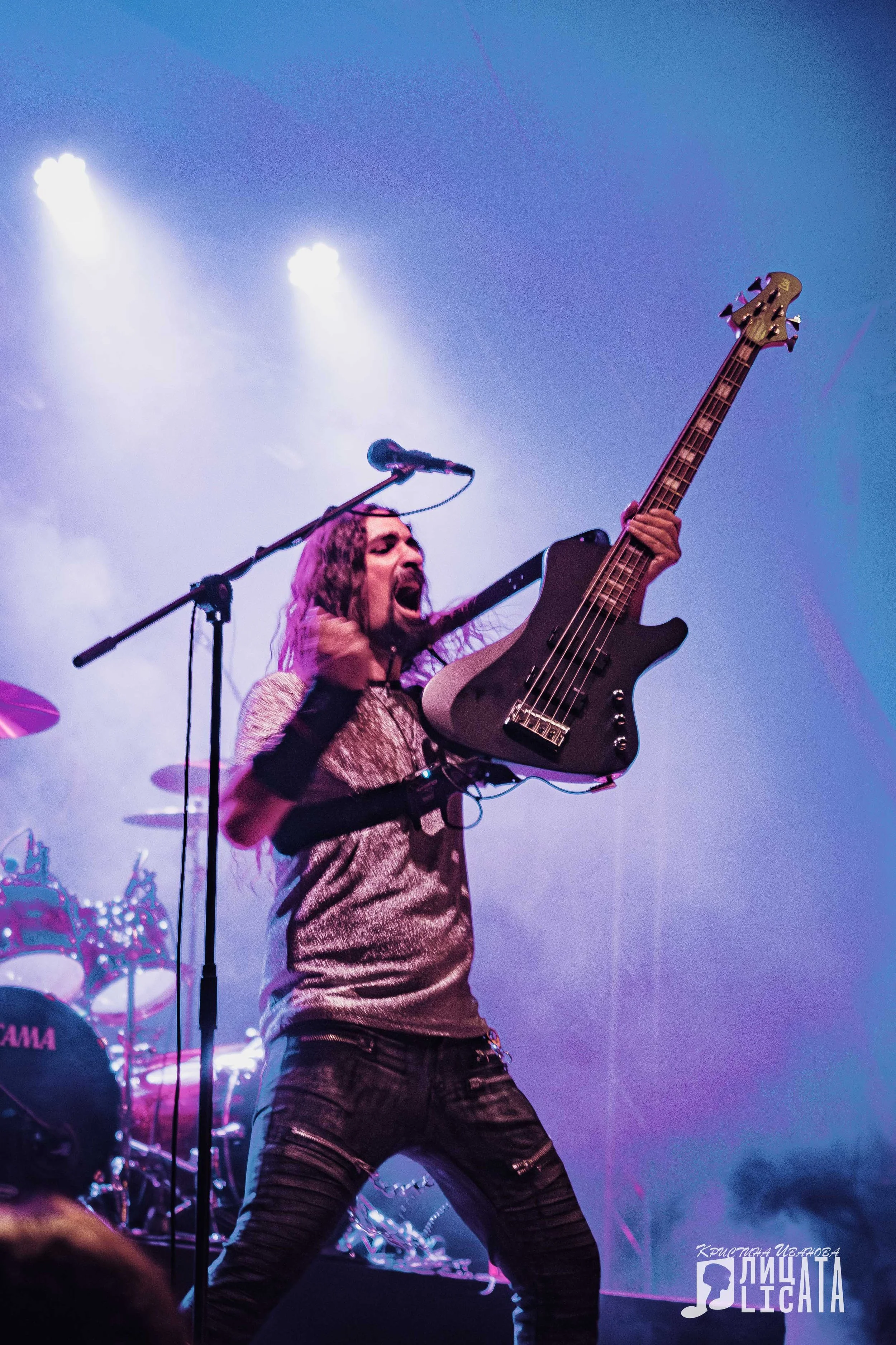 A male guitarist passionately performing on stage, holding an electric bass guitar, with purple stage lighting and drums in the background.