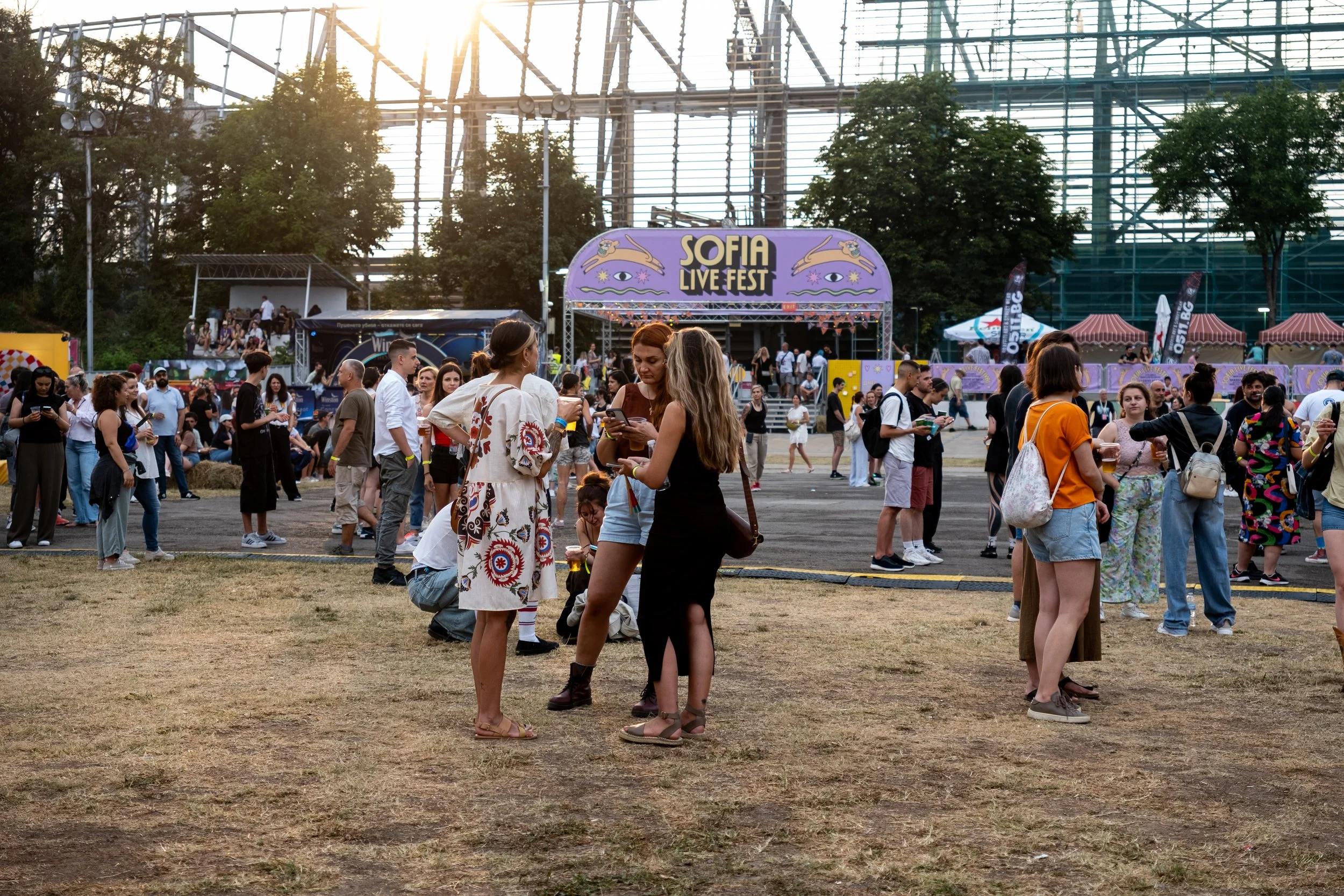 People attending Sofia Live Fest outdoor music event, with a stage in the background and festival tents on the right, during the late afternoon or early evening.