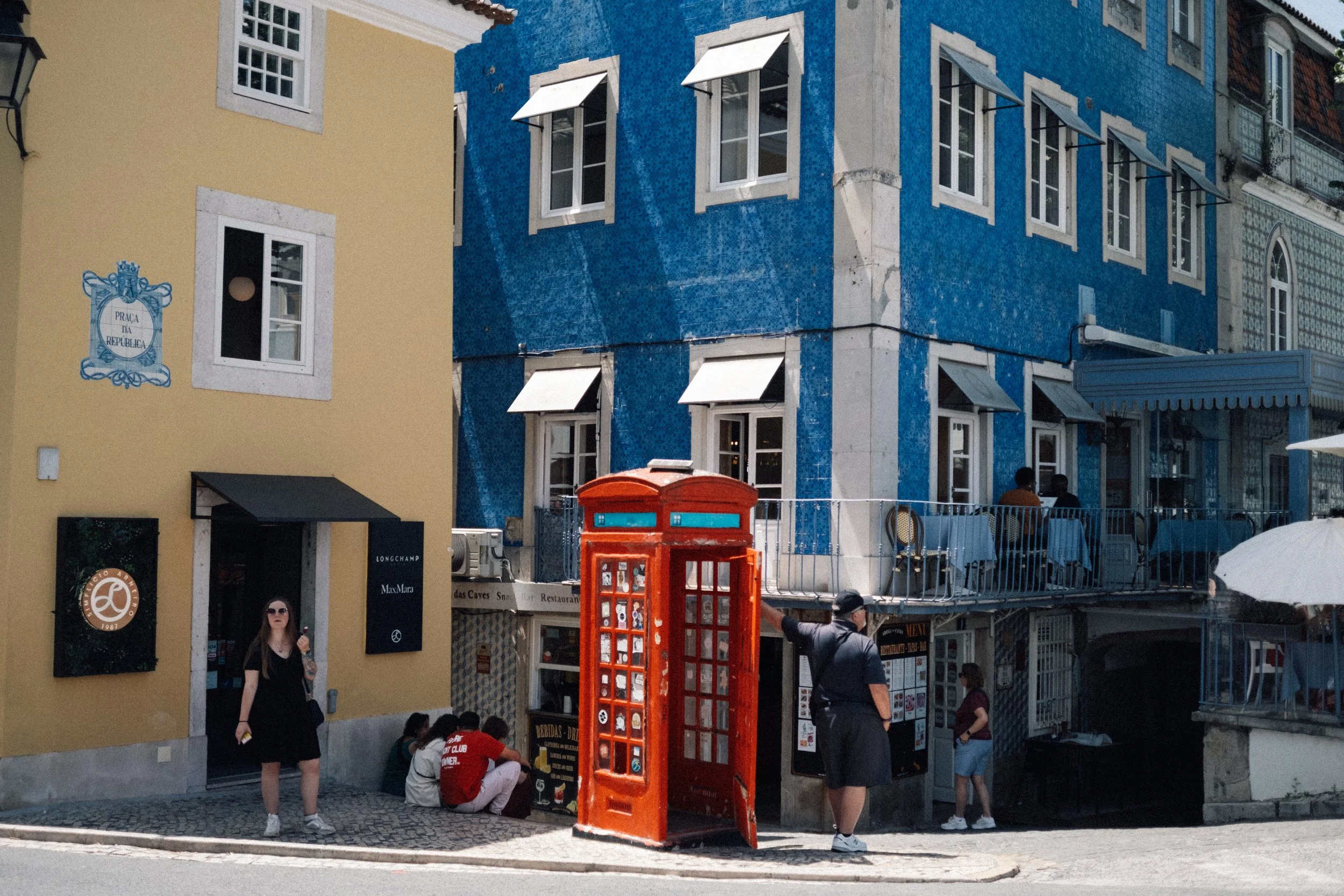 A colorful street scene featuring a blue building with white-framed windows and a yellow building with a sign. An orange telephone booth is on the sidewalk, with people sitting and walking nearby.