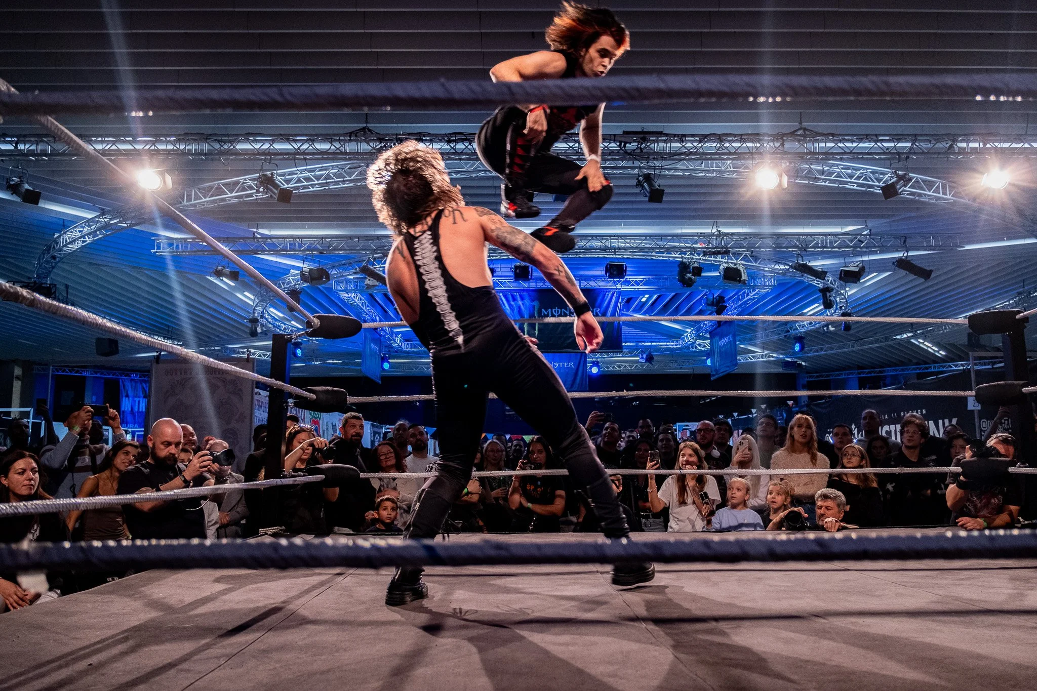 Two wrestlers are in the middle of a match in a wrestling ring, with a large audience watching. One wrestler is jumping toward the other from inside the ring, under bright overhead lights in an indoor arena.