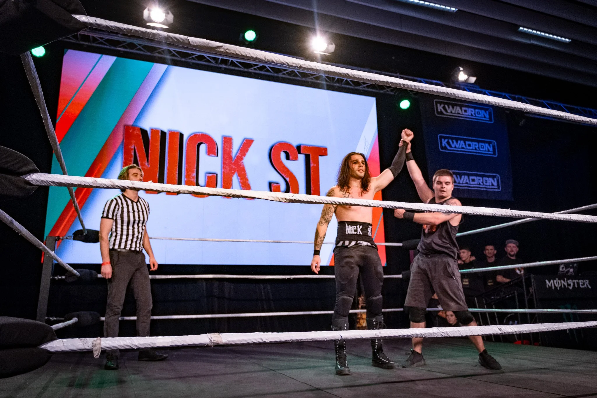 A professional wrestling match with two wrestlers in the ring and a referee observing. One wrestler is holding up the other's arm in victory while the audience watches.