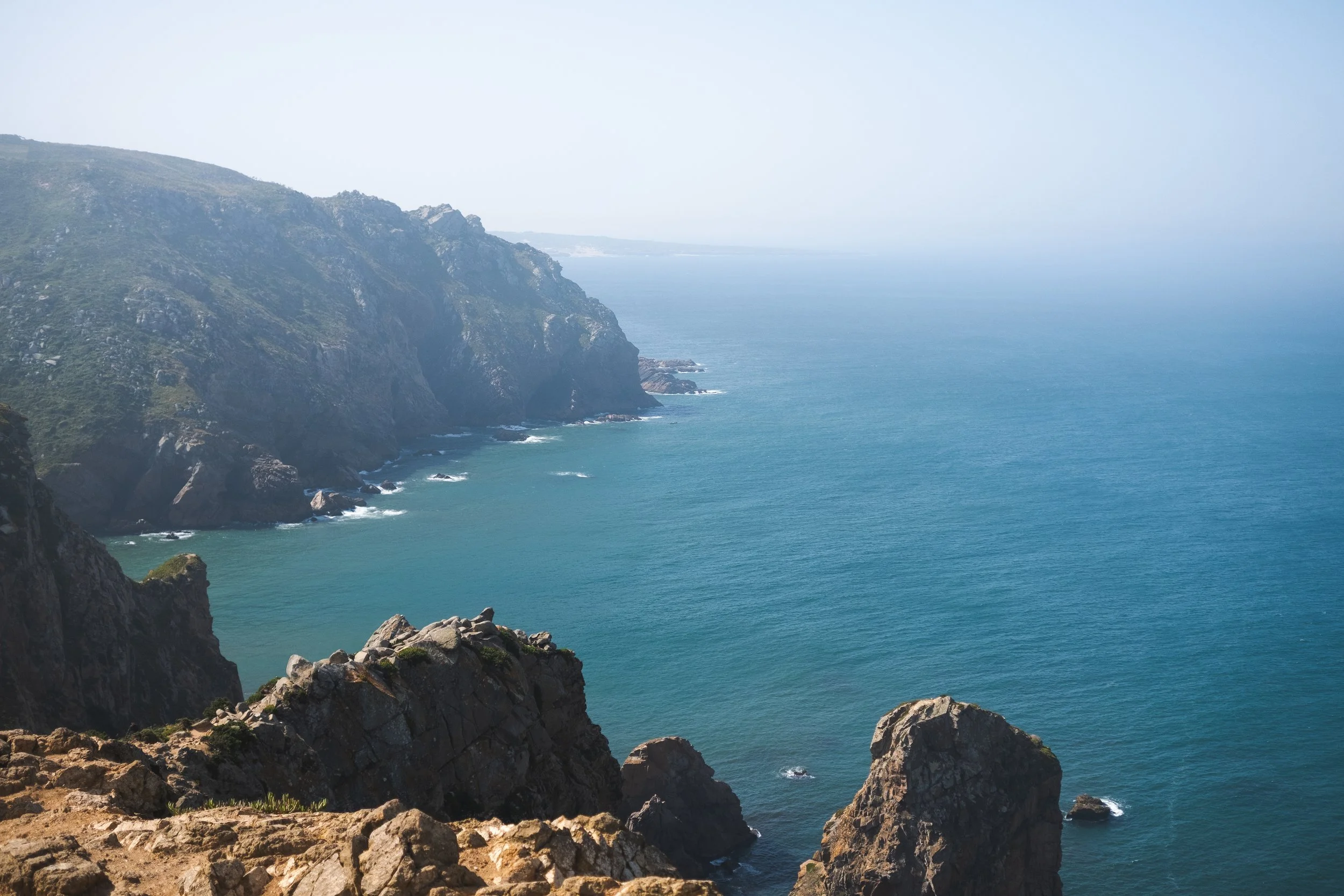 Coastal cliffs with rocky formations and the ocean, under a hazy sky.