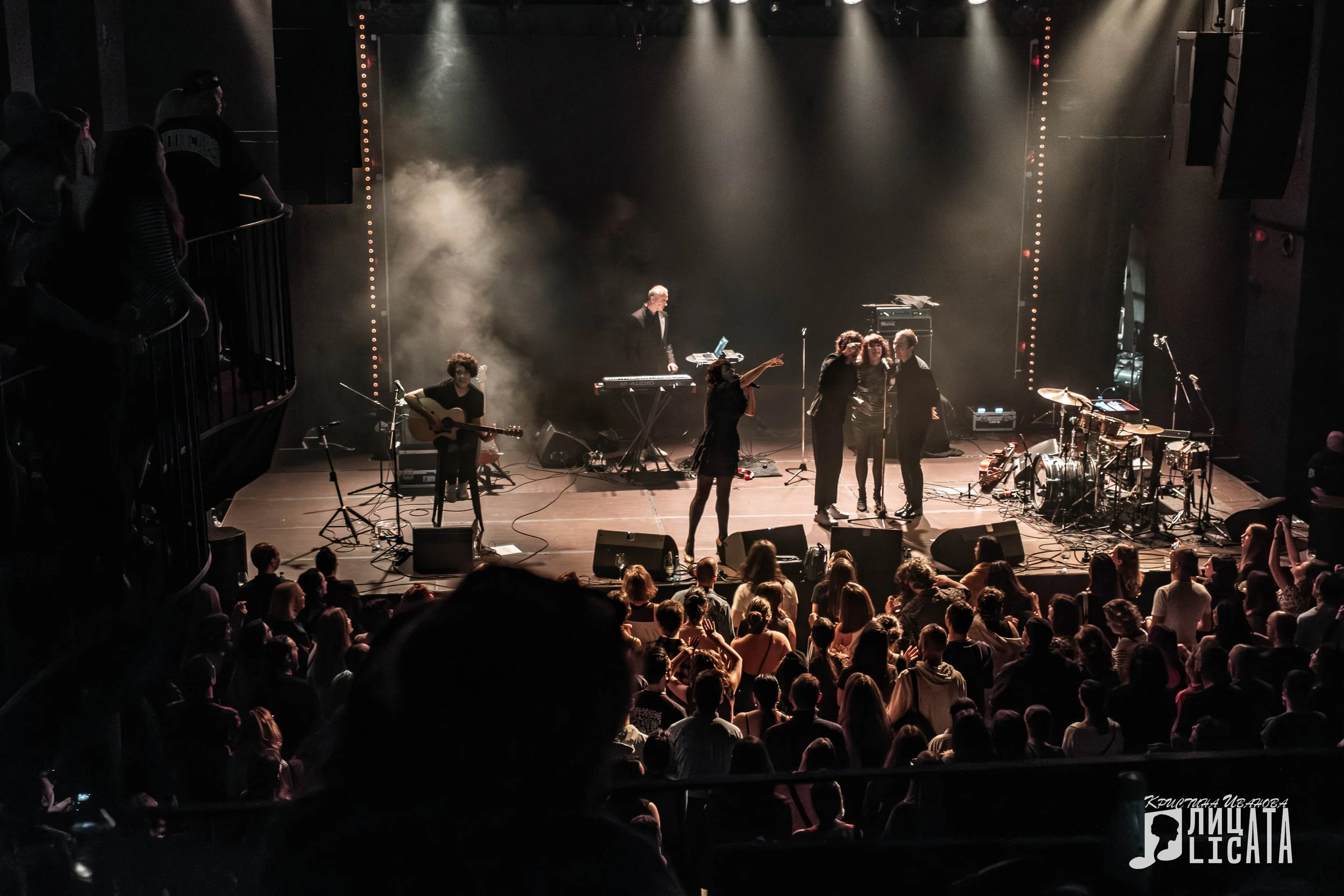 Nouvelle Vague French band on stage. Musicians on stage performing at a concert with an audience watching, stage lights, and musical instruments including guitars, drums, and keyboards.
