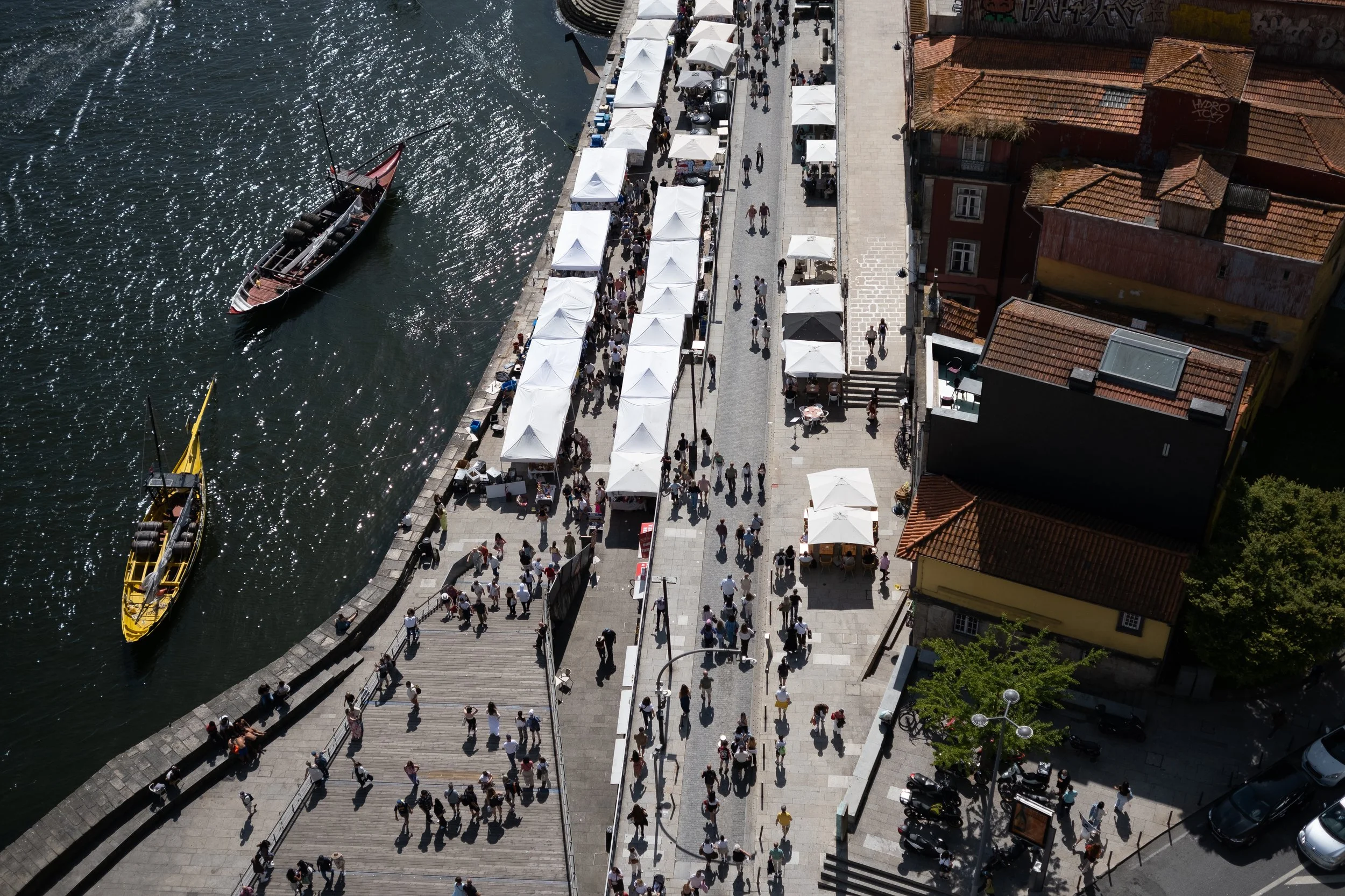 Aerial view of a riverside walkway with outdoor market stalls, boats docked along the water, and people walking along the street and stairs.