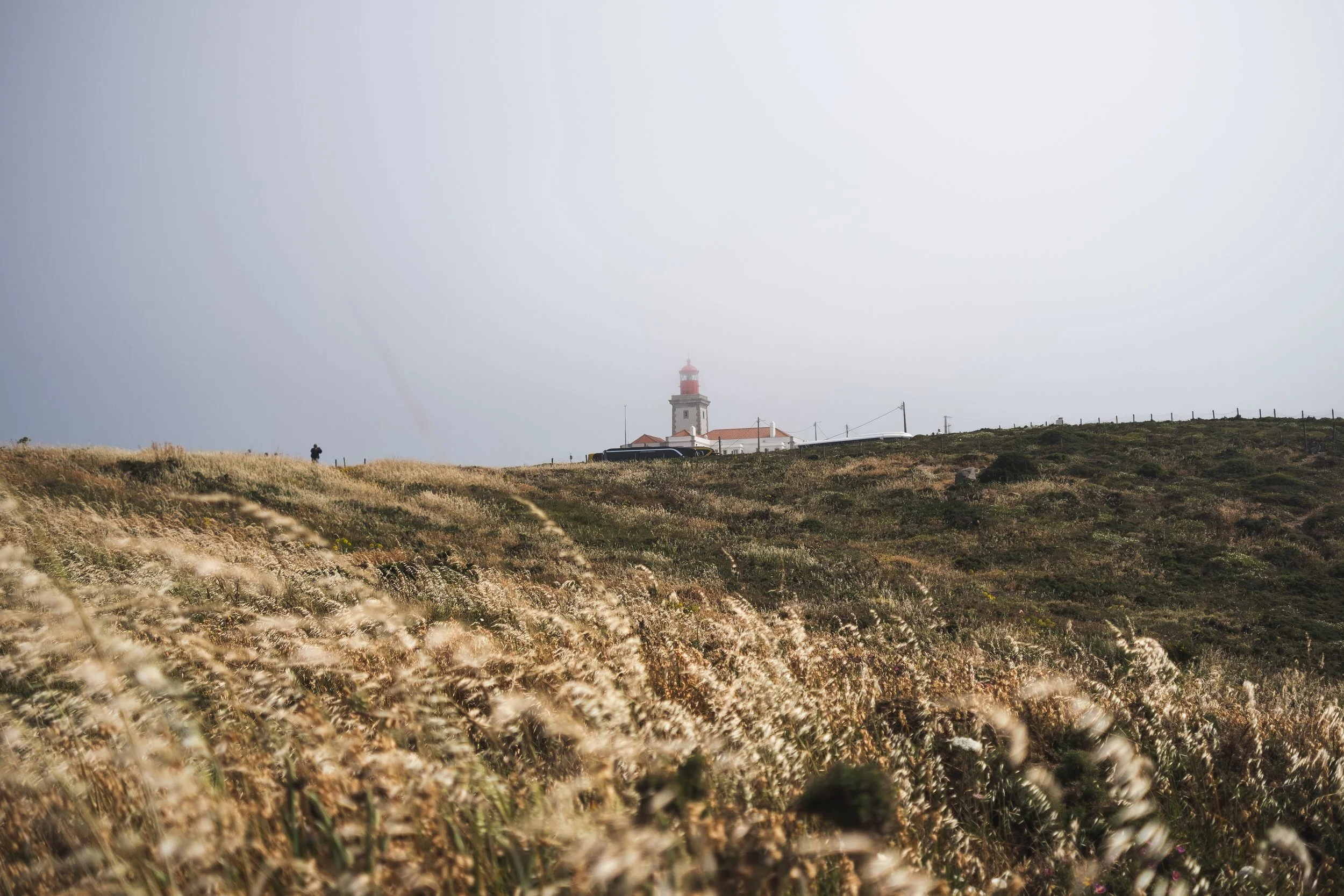 A lighthouse on a hilltop with a person walking nearby, cloudy sky, and tall grass in the foreground.