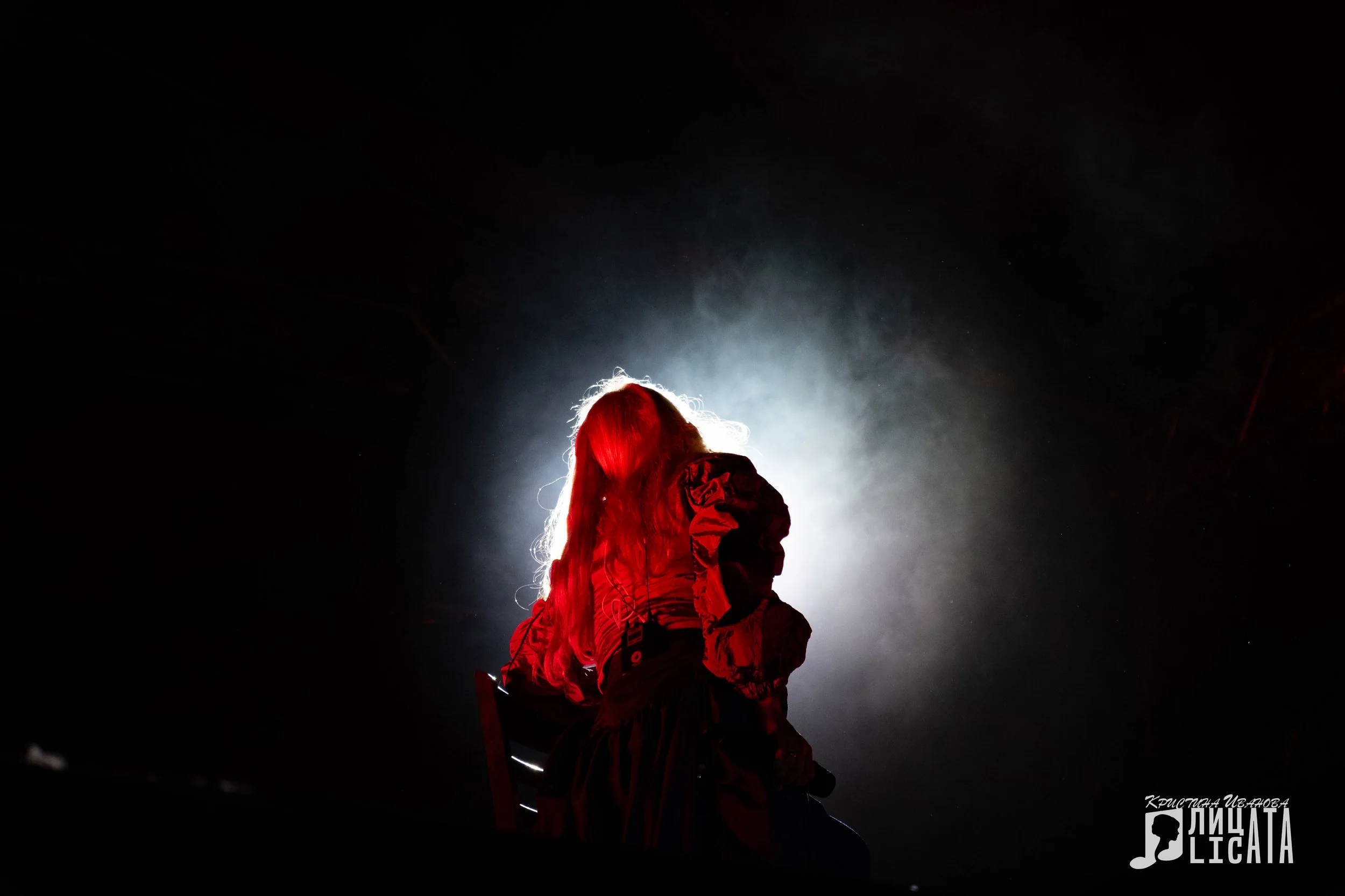 A person with long hair sitting on a chair in a dark room, illuminated from behind with a bright light creating a silhouette effect.