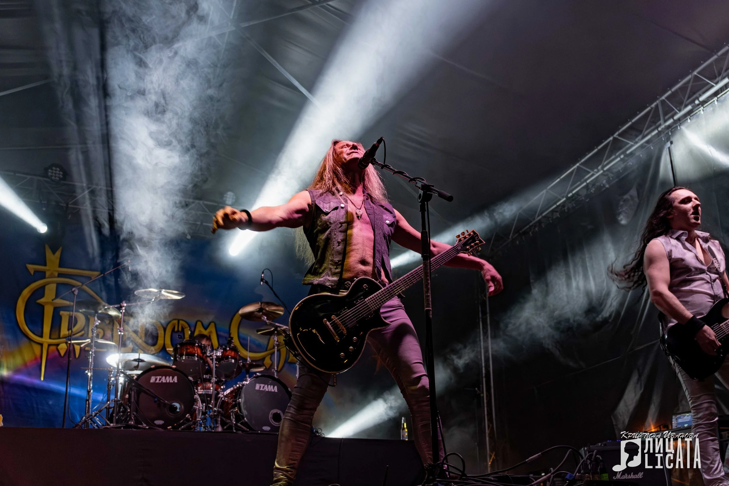 freedom Call band performing on stage with smoke and lights, featuring a long-haired male guitarist singing into a microphone with his arms outstretched, and another band member to the right also playing guitar.