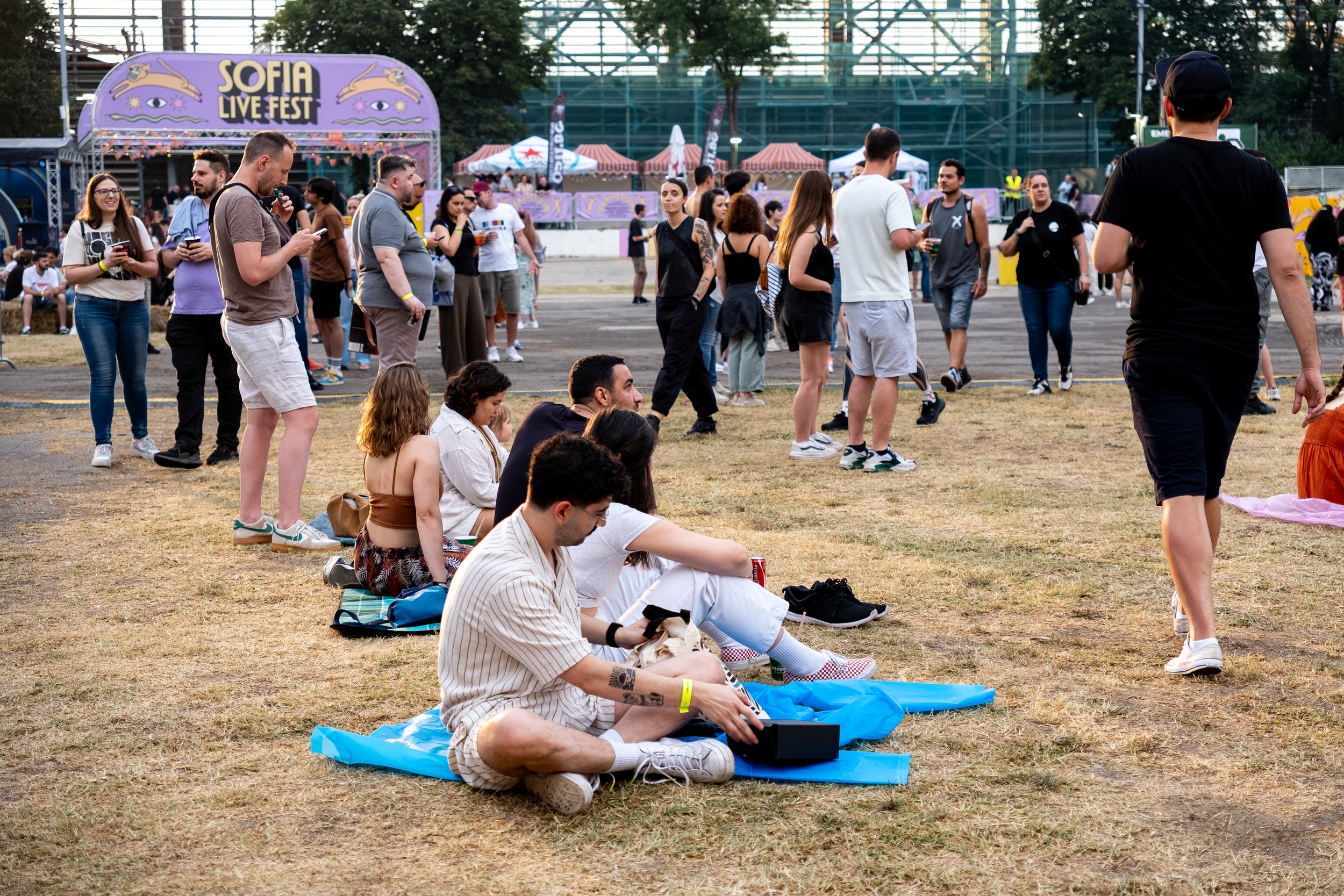 People attending Sofia Live Fest outdoor concert, some sitting on the grass, others standing or walking near the stage with purple signage, tents, and trees in the background.