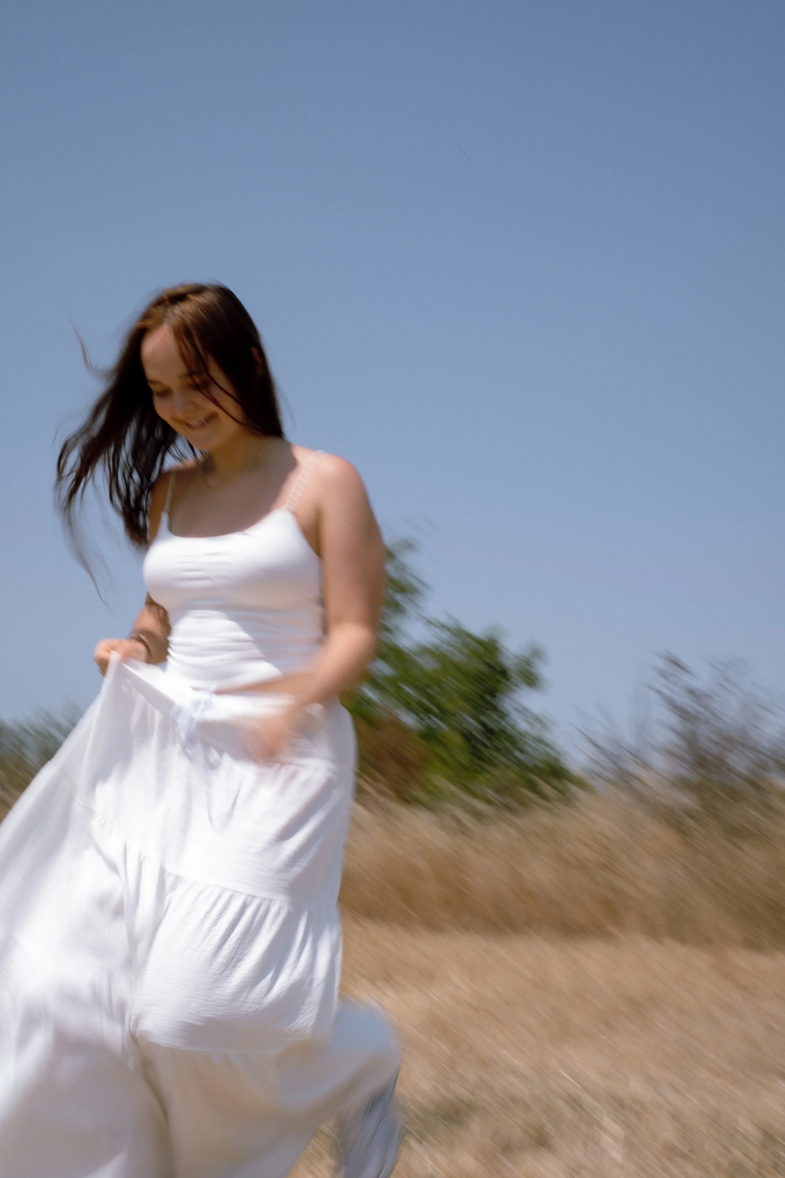 A woman in a white dress smiling and holding the hem of her dress while running outdoors during the daytime with a blue sky and dry grassy field in the background.