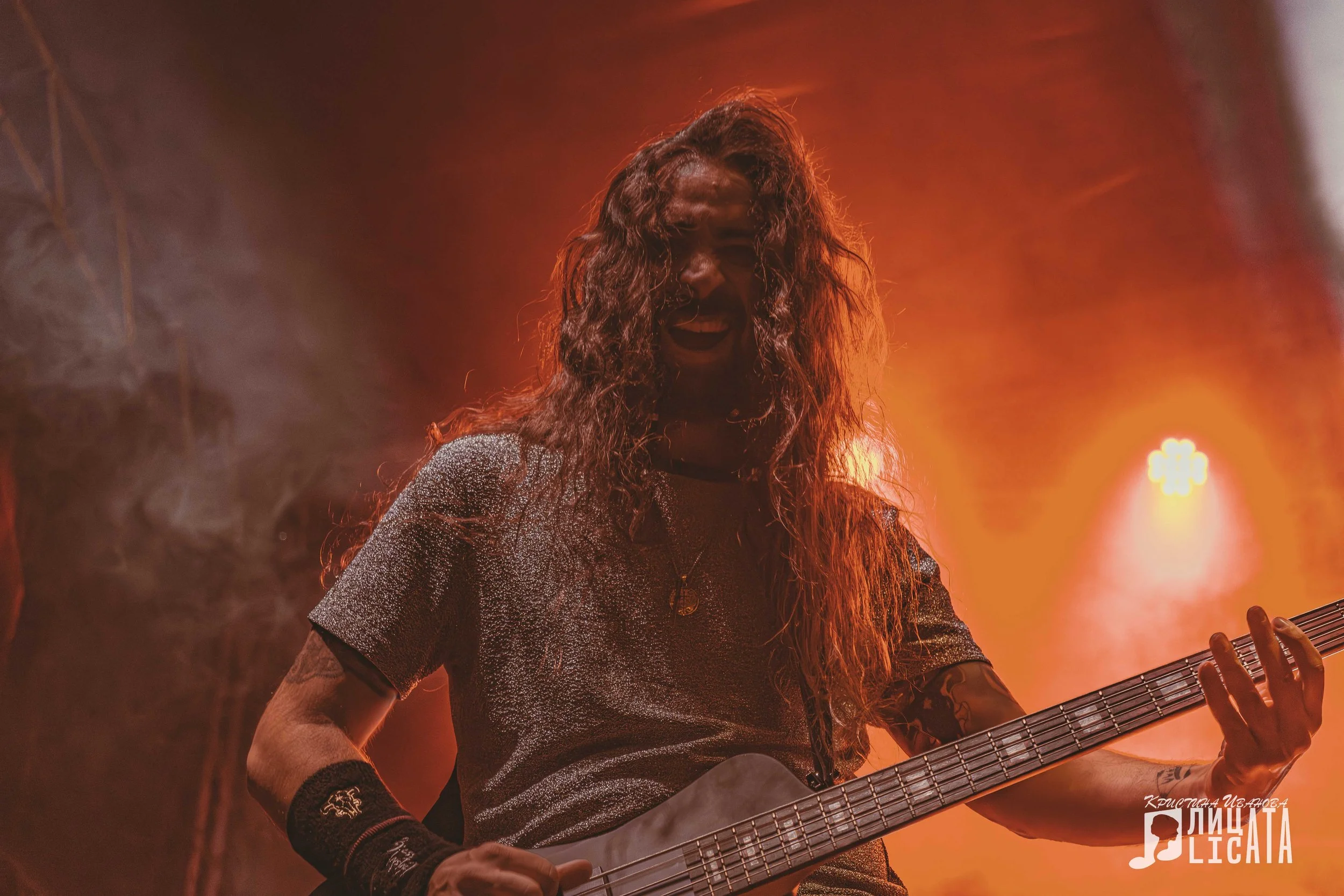 A male guitarist passionately performing on stage, holding an electric bass guitar, with yellow stage lighting and drums in the background.