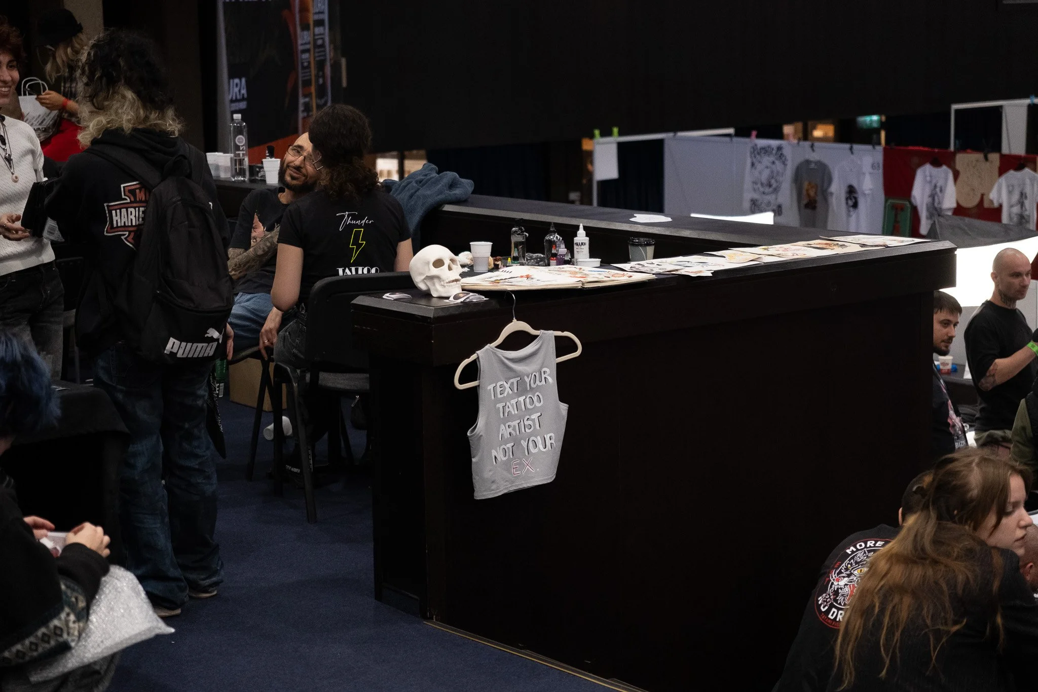 Tattoo booth with a skull on the counter, a tank top hanging with the text 'TEXT YOUR TATTOO ARTIST NOT YOUR EX,' and people engaging in conversation at a tattoo convention.
