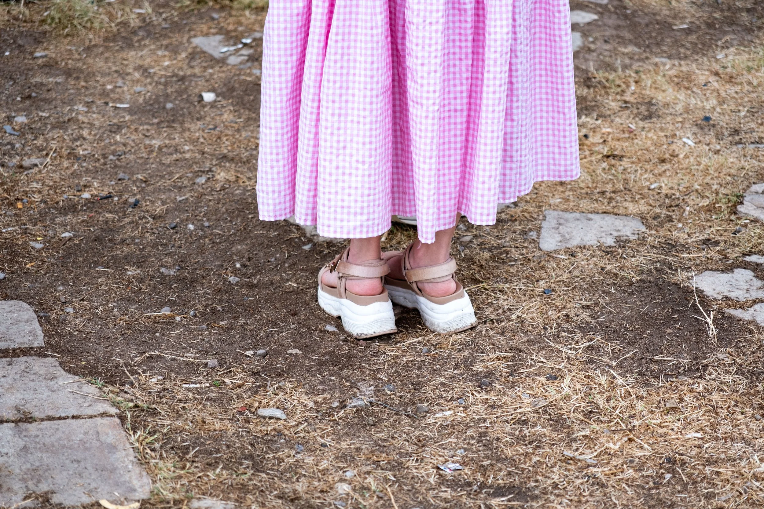 Person wearing a pink gingham dress and platform sandals standing on dirt and stone pathway.