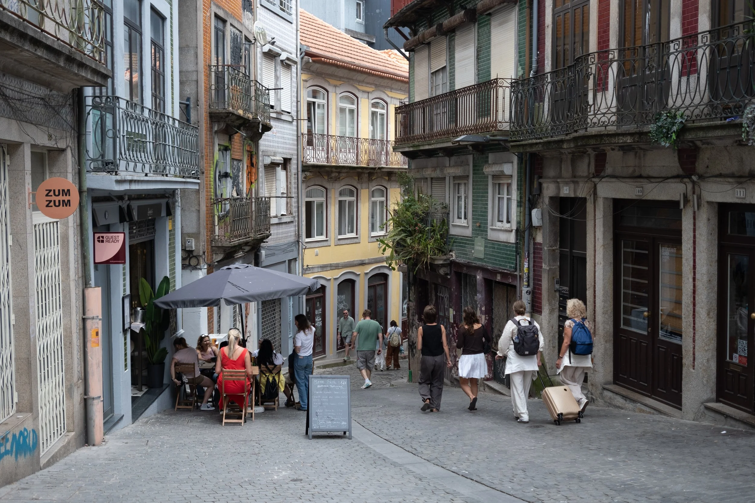 A narrow European street with colorful old buildings, some with balconies, with people walking and a small outdoor café having customers, and signs for 'ZUM ZUM' and a chalkboard menu.