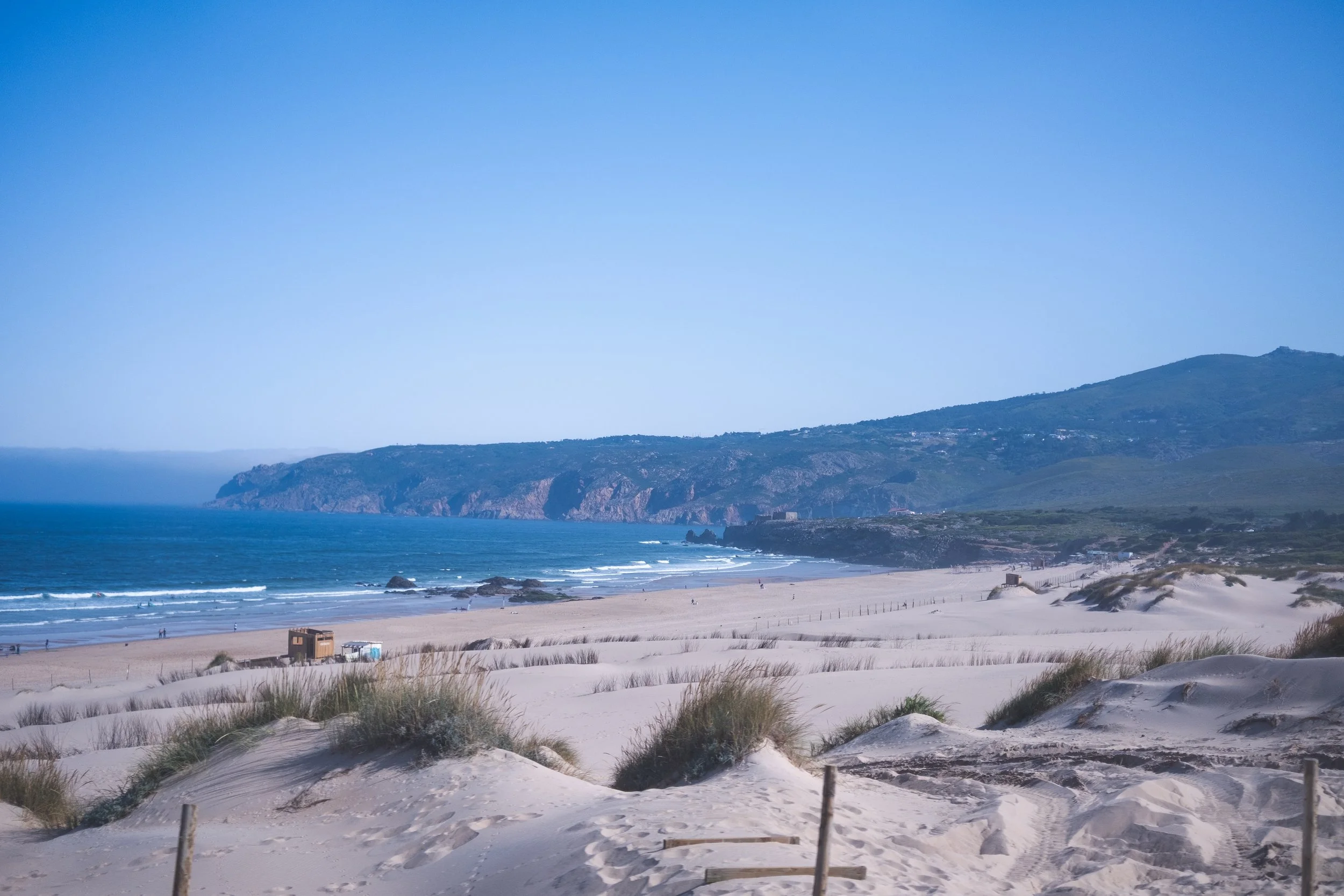 Sandy beach with dunes and grass, shoreline with gentle waves, rocky cliffs in the distance under a clear blue sky.