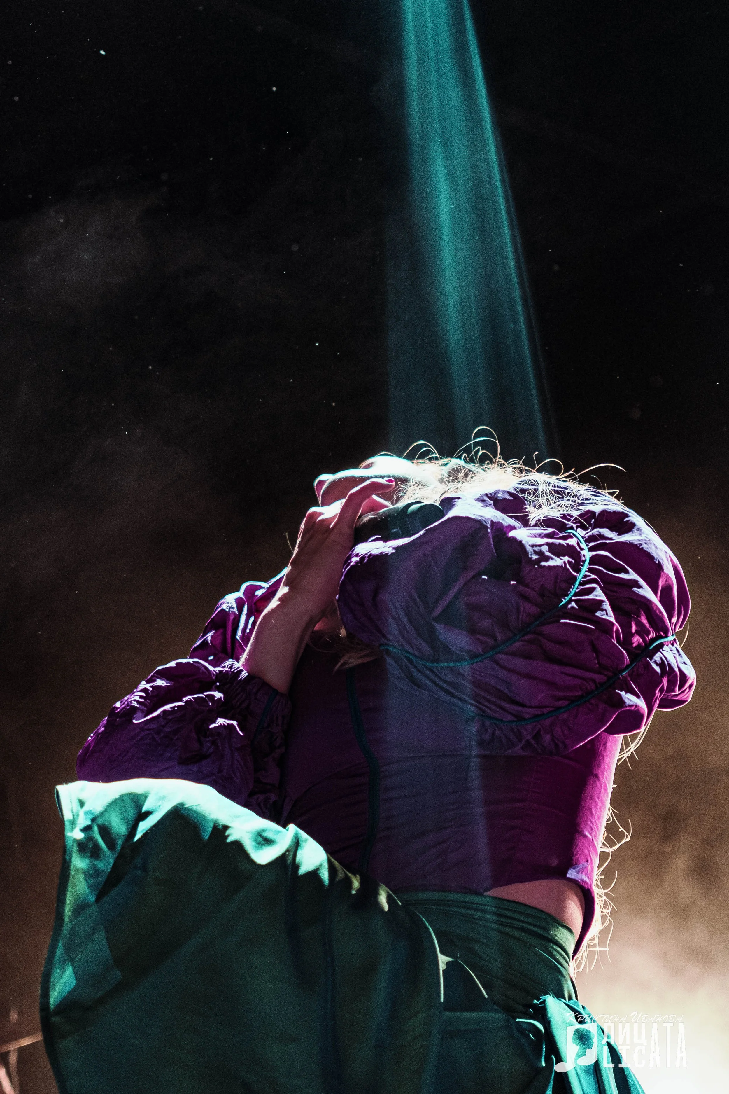 Person wearing a purple jacket and blue pants, holding a headlamp to their head, standing outdoors at night with a starry sky and a beam of light in the background.