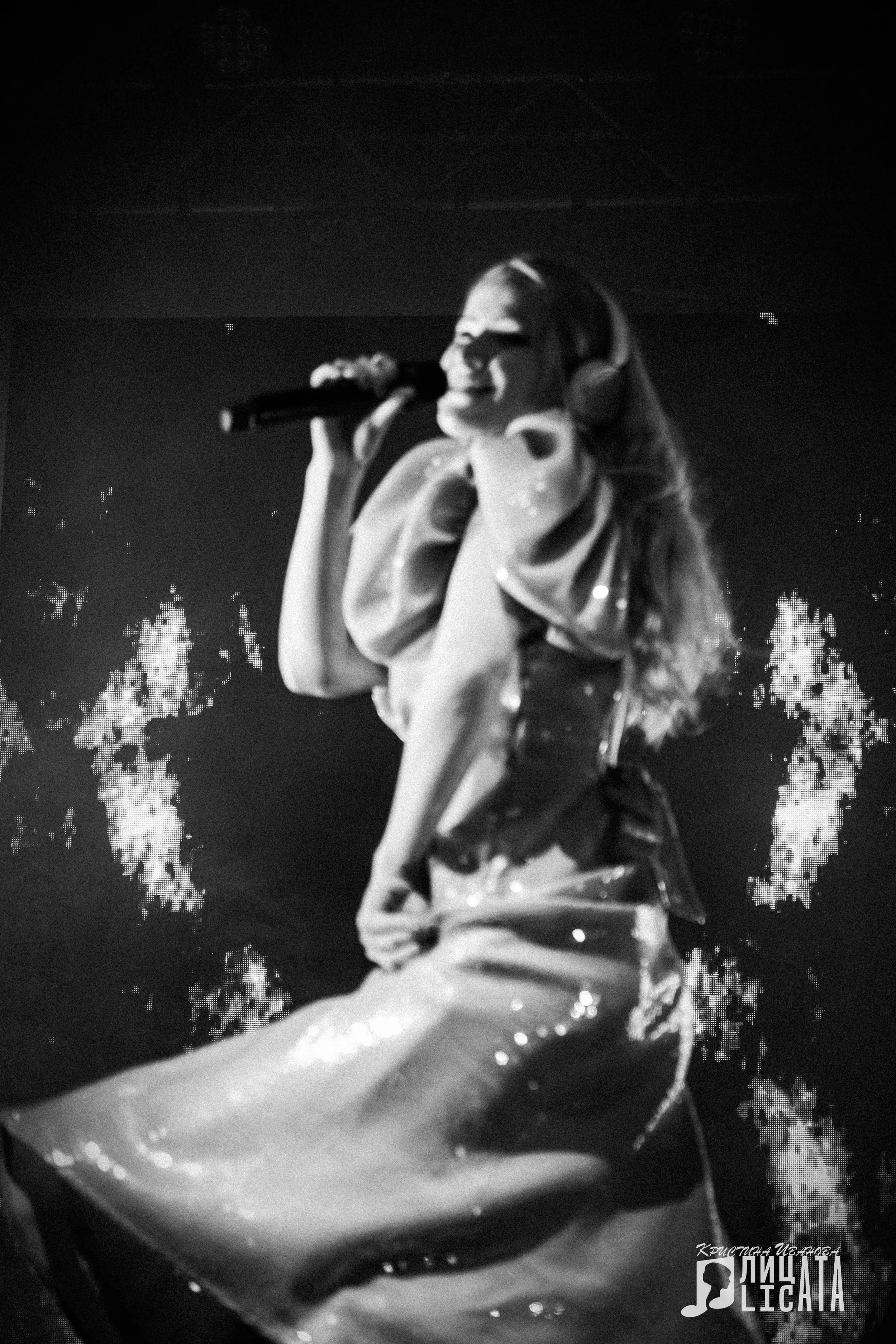 A woman performing on stage, singing into a microphone, wearing large headphones and a shiny outfit, with a dark background and bright lights.