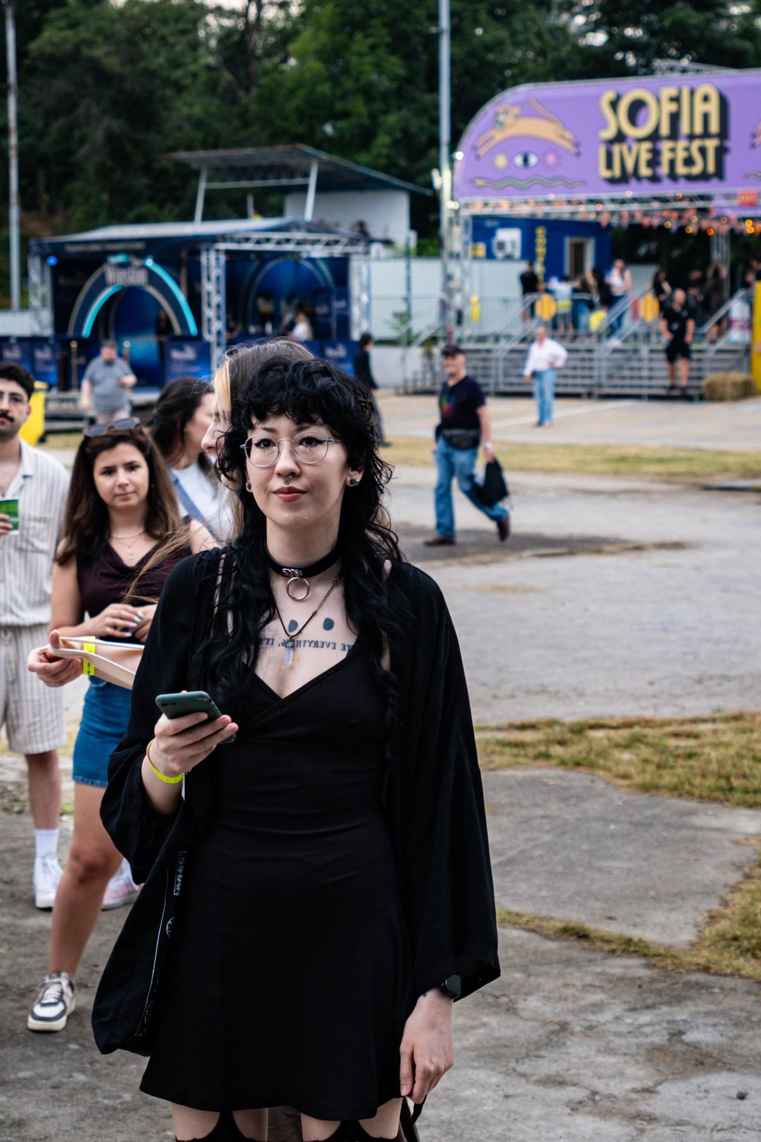 A young woman with black curly hair and glasses standing at a festival with a stage in the background. She is wearing black clothing and holding a smartphone.