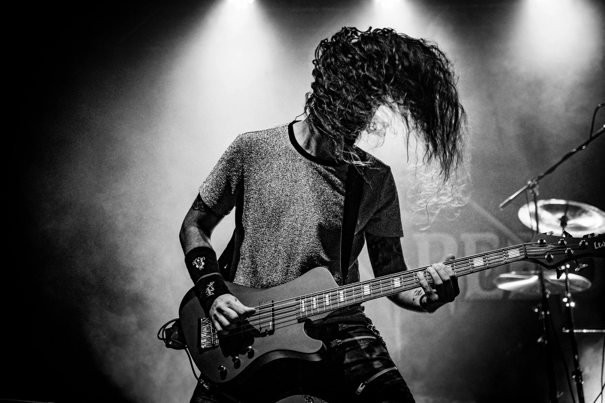 A rock musician playing an electric guitar on stage, with long hair covering their face and wearing a short-sleeved shirt, in black and white.