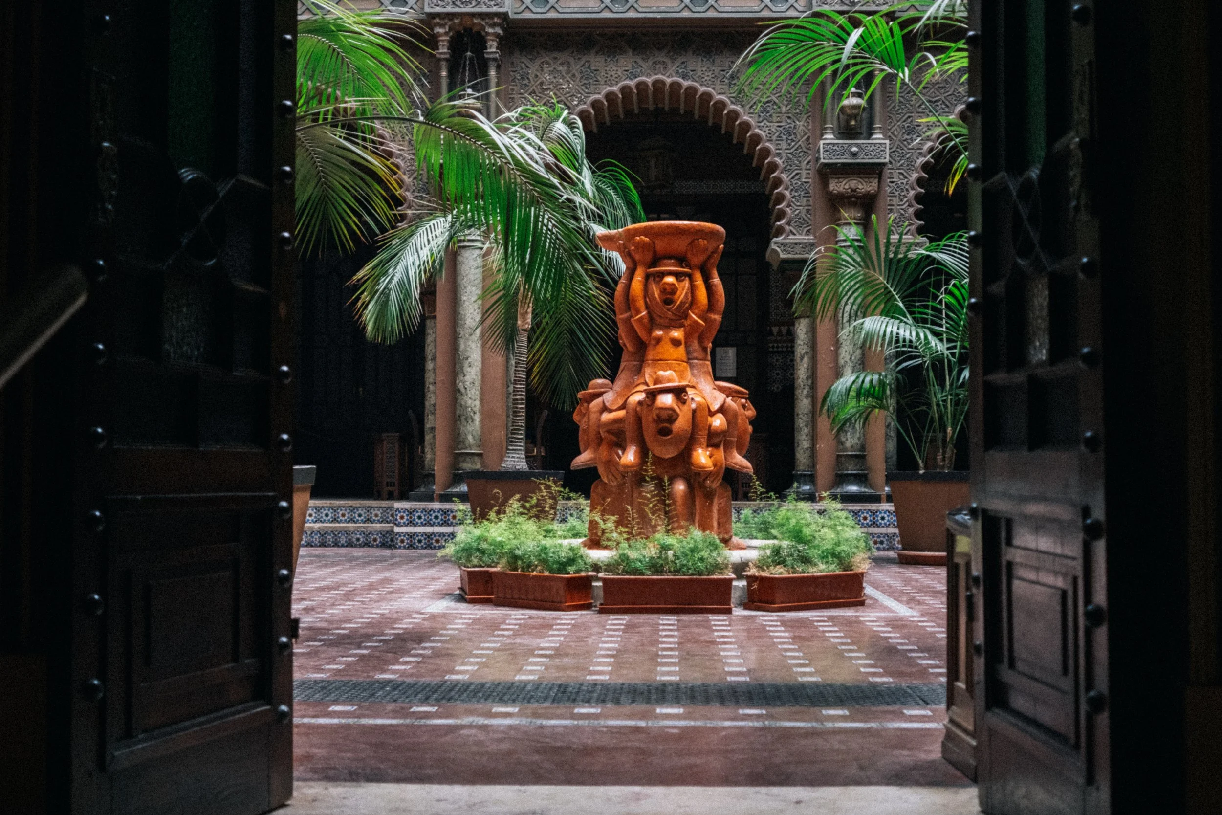View through open dark wooden doors revealing an indoor courtyard with a central terracotta sculpture of mythological figures, surrounded by potted palms and decorative tiled flooring in a historical architectural setting.