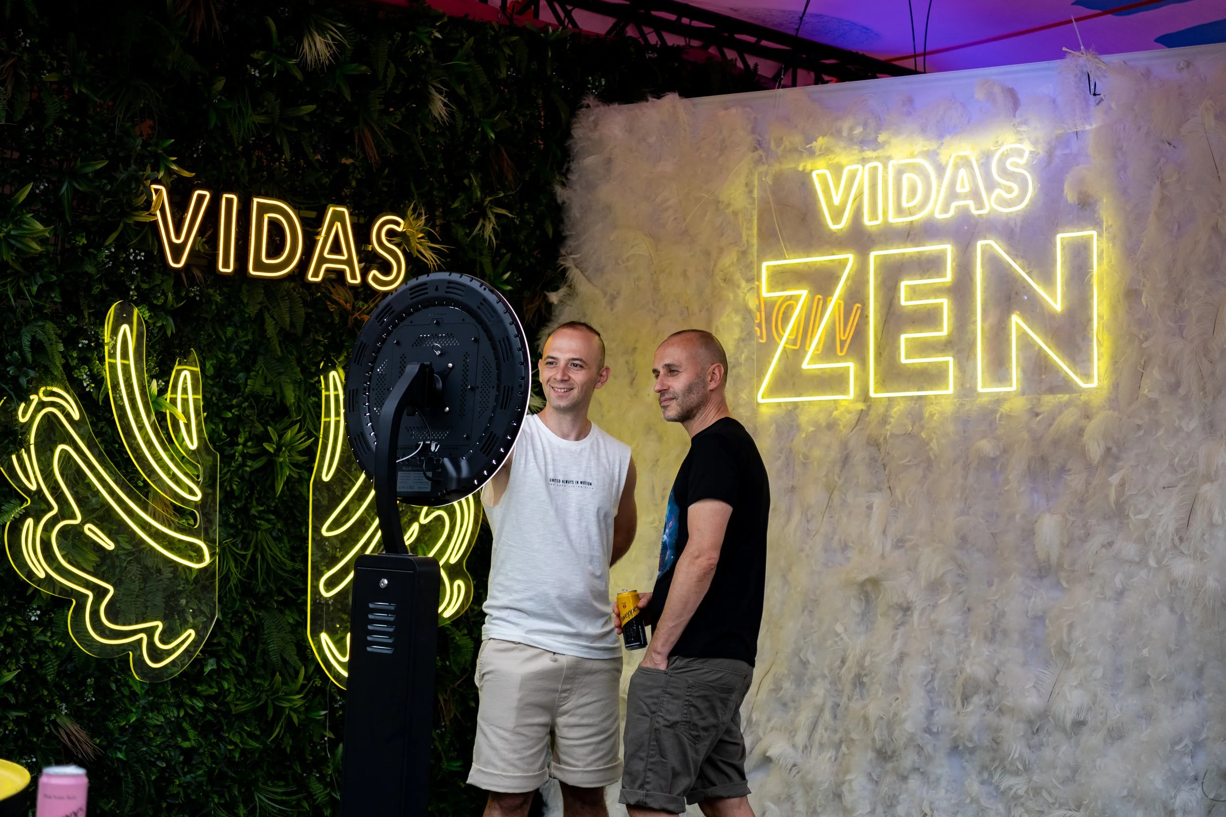 Two men in casual clothing are standing near a fan, smiling and talking at a party with neon signs and greenery. One person is holding a beverage can. The background features neon signs that read 'VIDAS' and 'VIDAS ZEN,' with yellow butterfly-shaped 