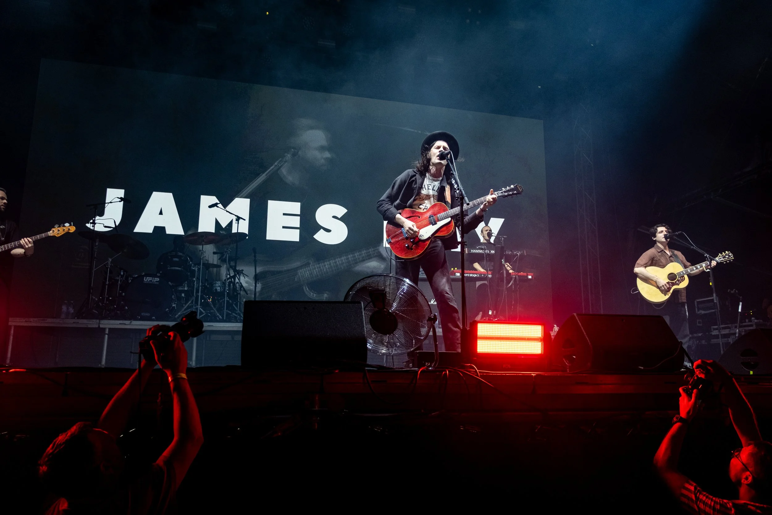 Concert Photographer captured James Bay on stage playing guitar and singing with a large screen behind them displaying the name 'JAMES'. Band members play instruments in the background, and photographers capture the performance in the foreground.