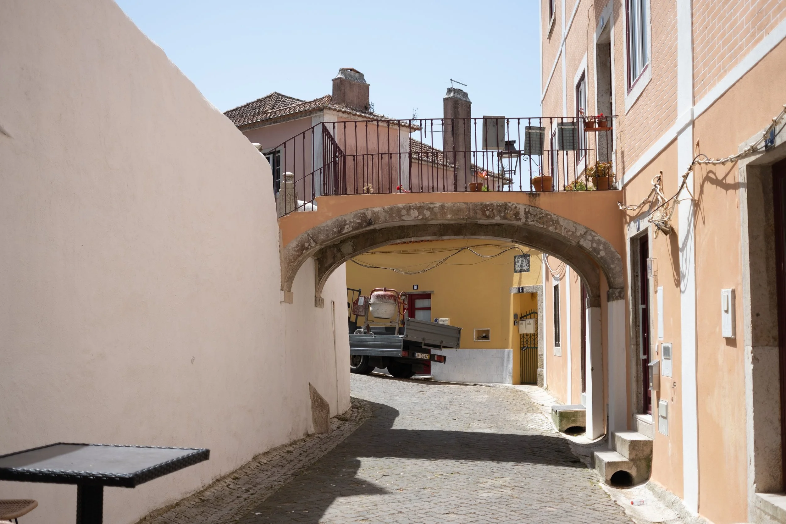 Narrow cobblestone street with a stone archway and colorful buildings on either side. A small truck is parked in the background, and potted plants are on the balcony above.