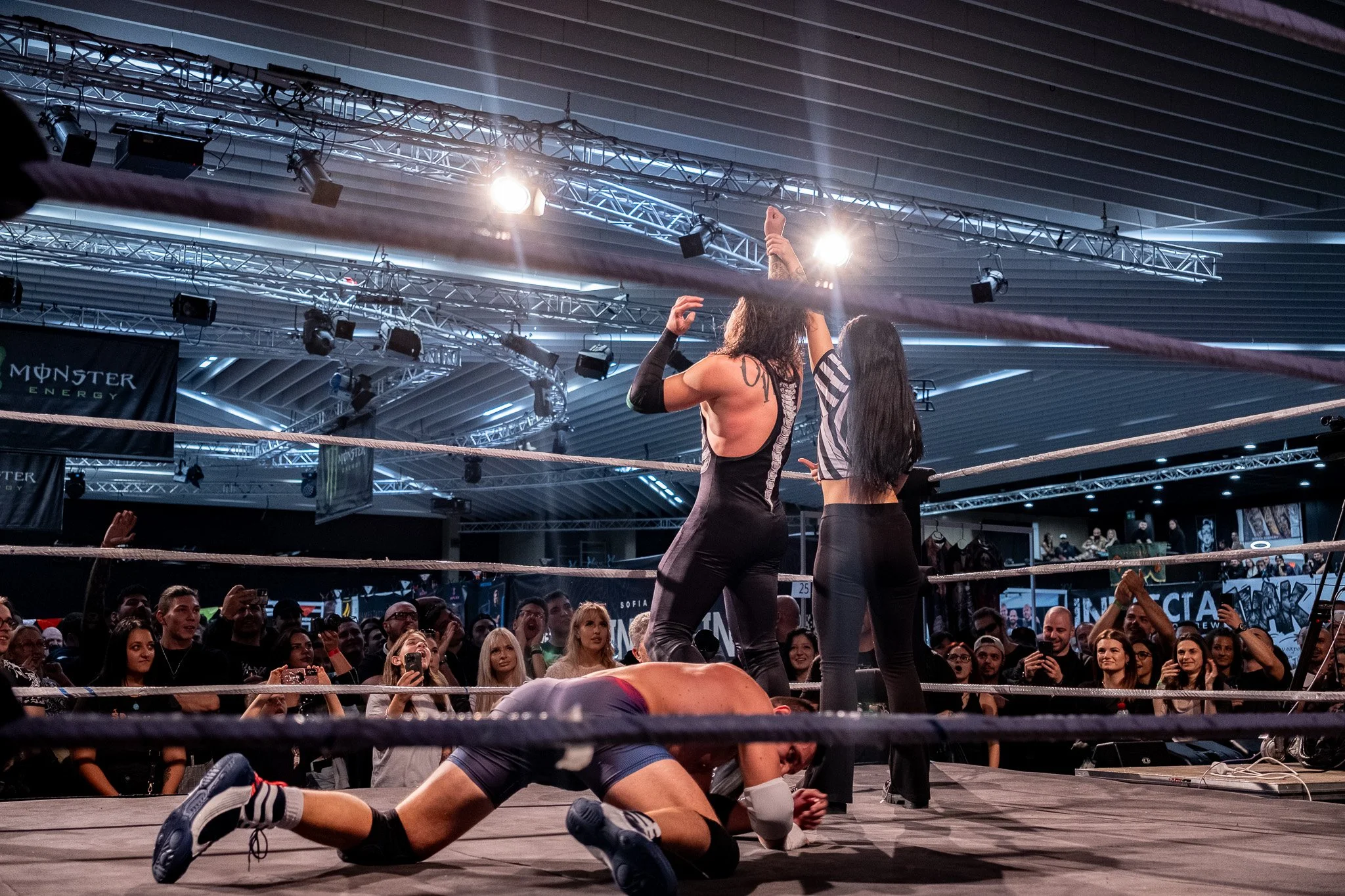 A wrestling match in a crowded indoor arena, with a wrestler on the ground and two female wrestlers standing inside the ring, one raising her arm in victory and the other celebrating. The audience watches enthusiastically.