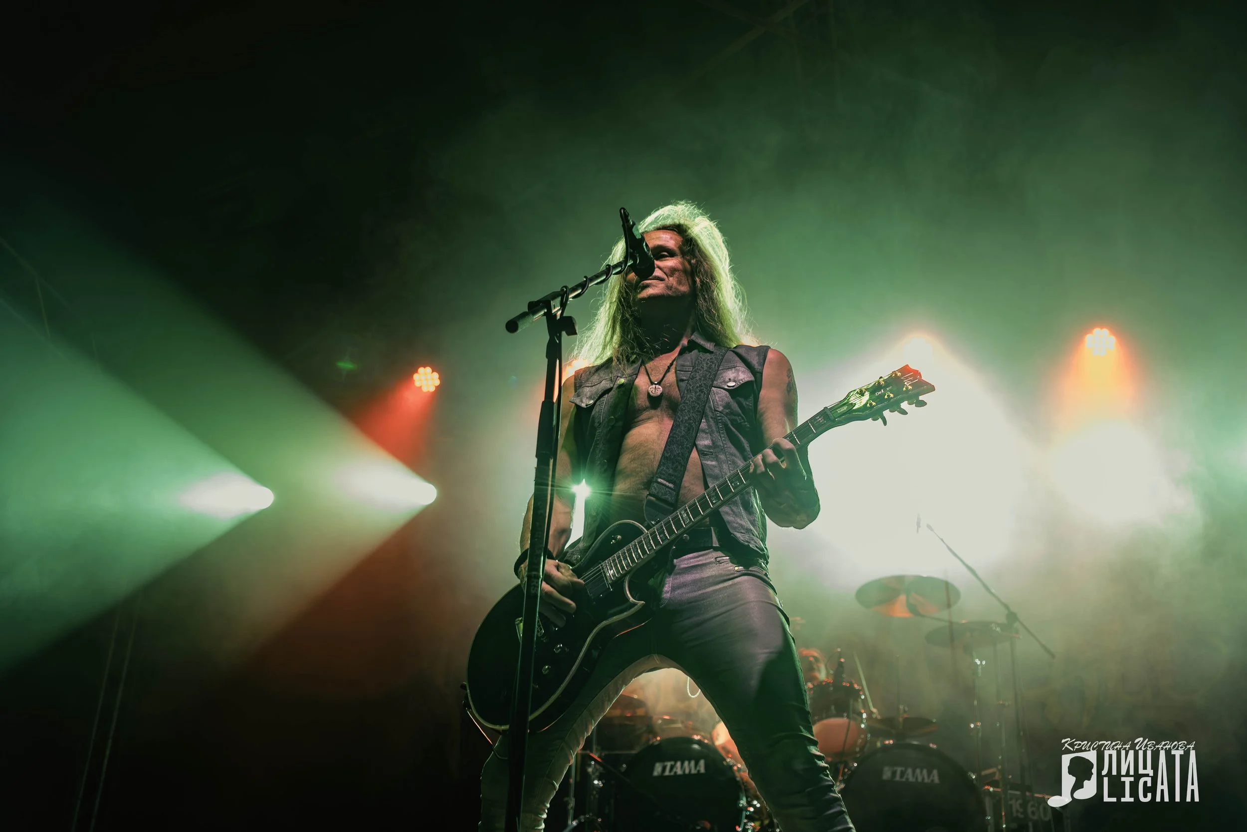 Freedom Call band.  Long-haired male guitarist singing into a microphone on stage, with drums in the background and stage lights creating a dramatic effect.