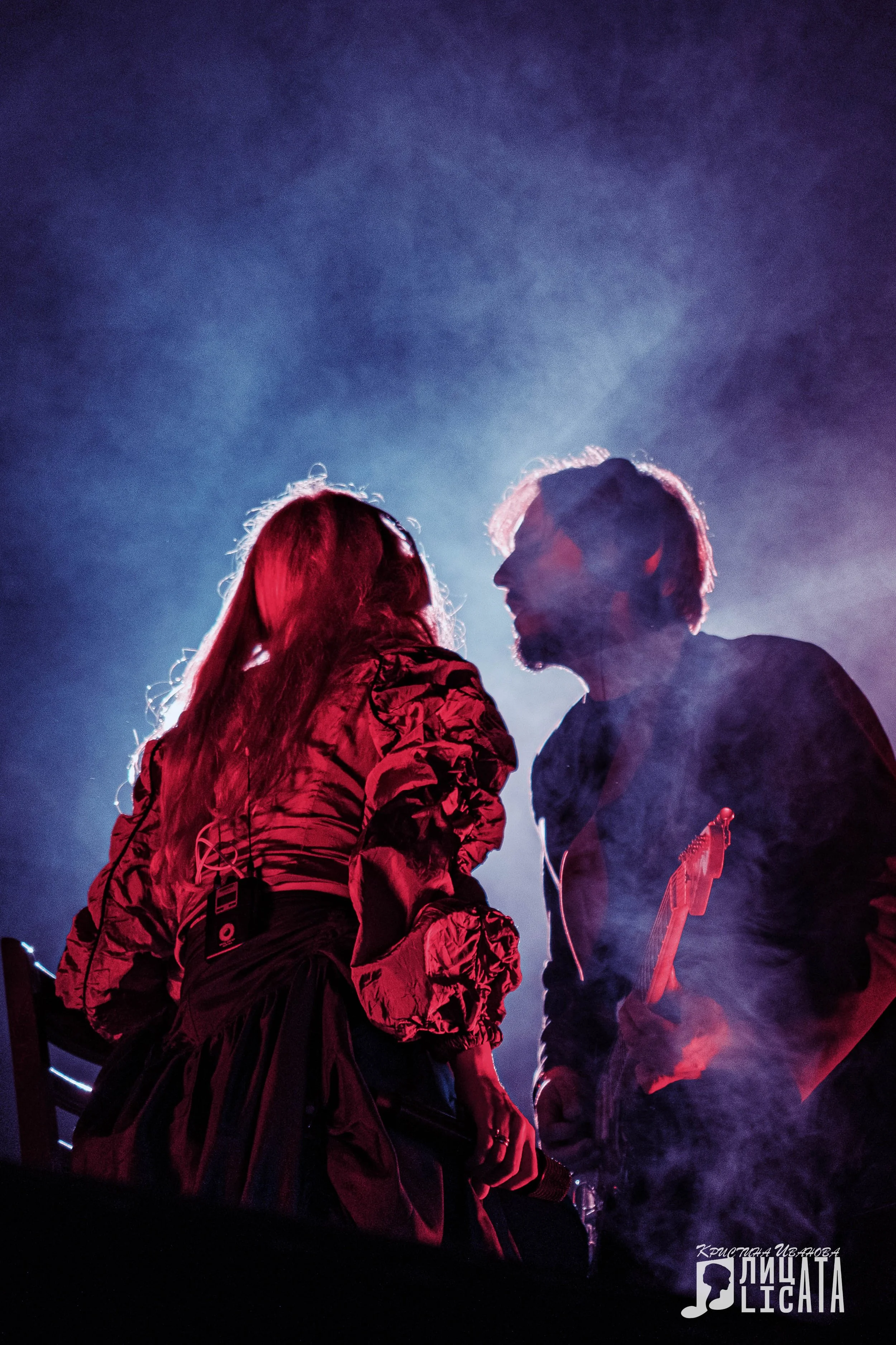 Two musicians, a woman with long hair and a man with headphones, face each other intensely on stage with dramatic backlighting and smoke.