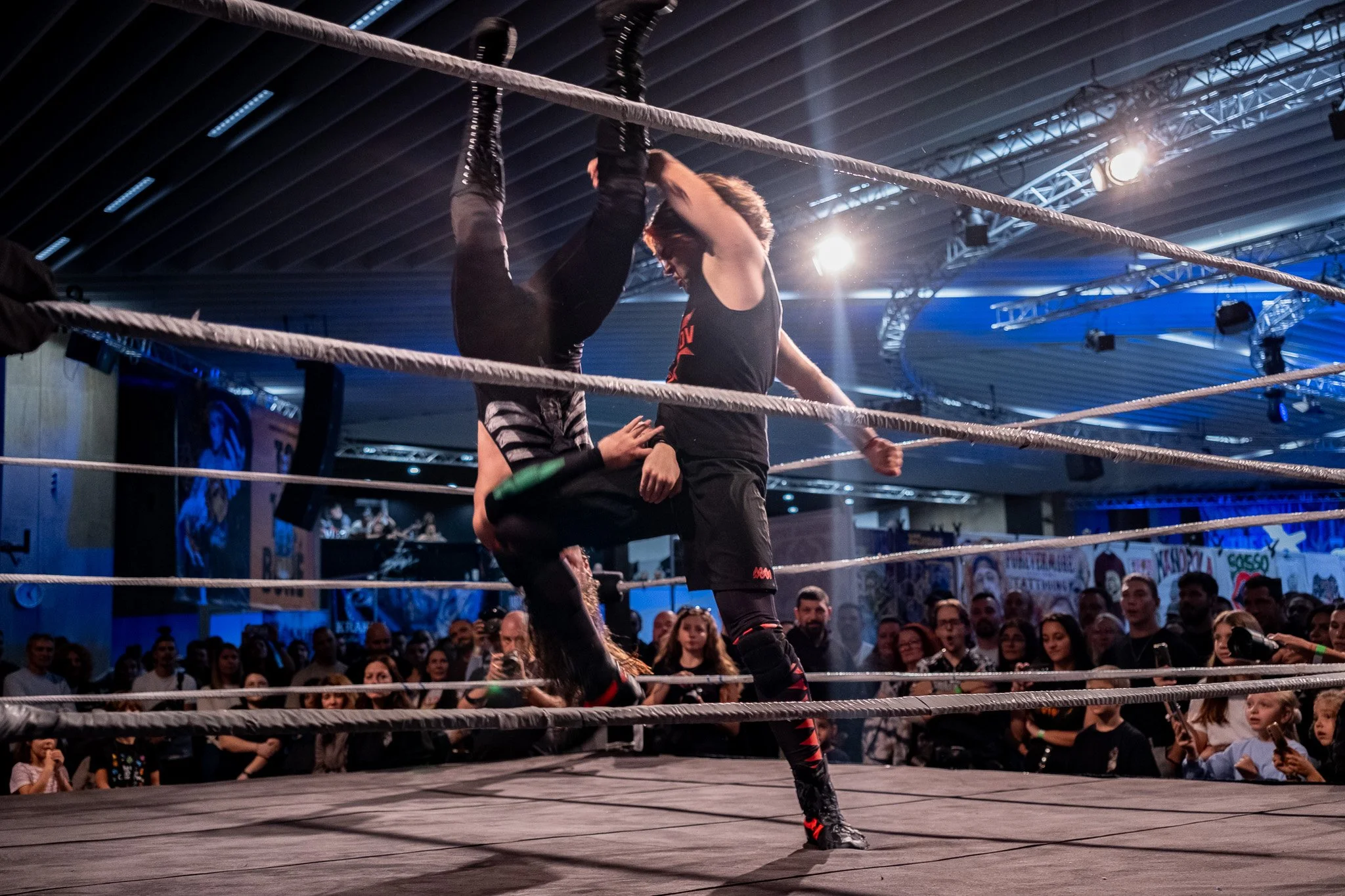 Two wrestlers engaged in a match inside a wrestling ring, with an audience watching in the background.