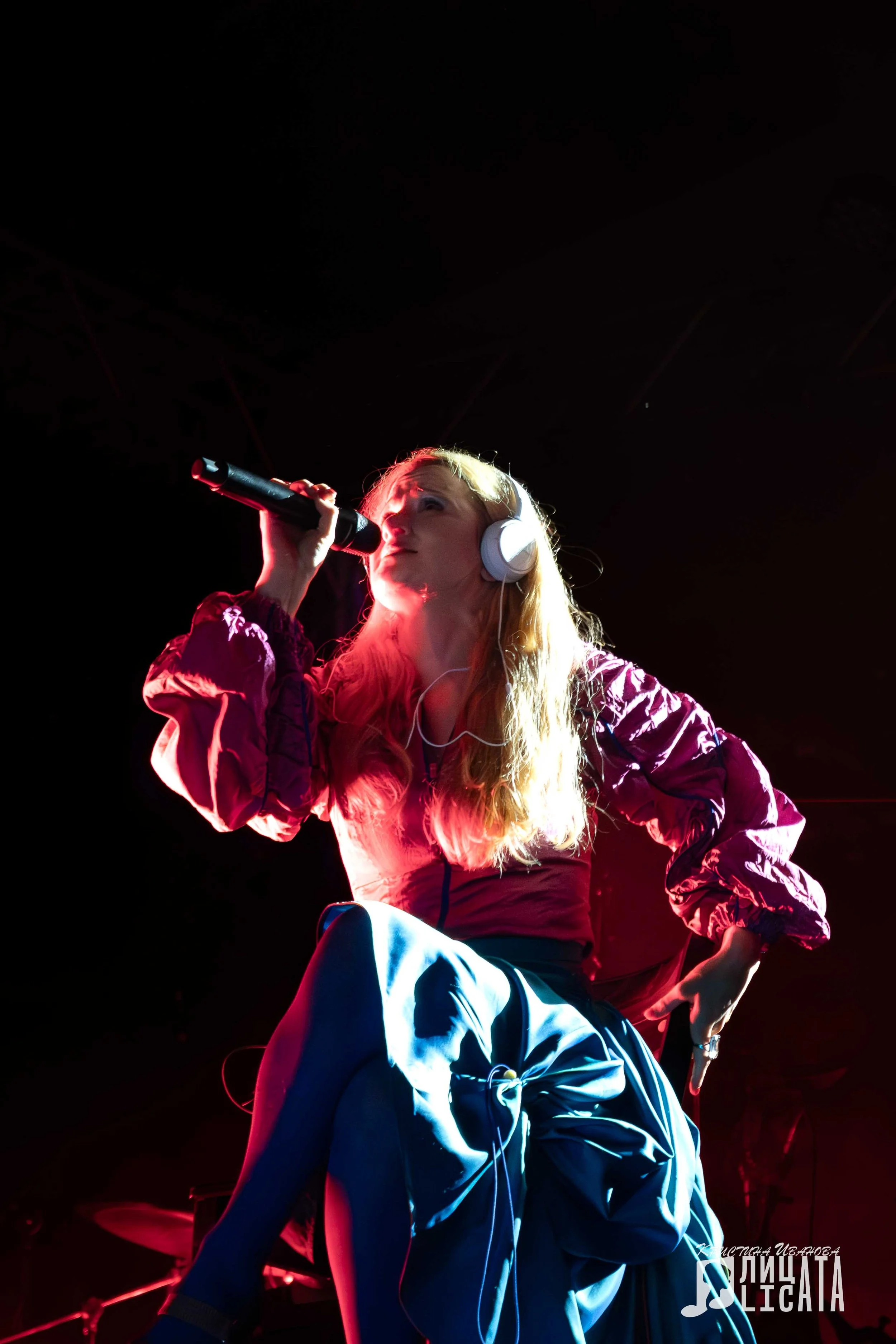 A woman with blonde hair wearing headphones, singing into a microphone, on stage with colorful lighting.