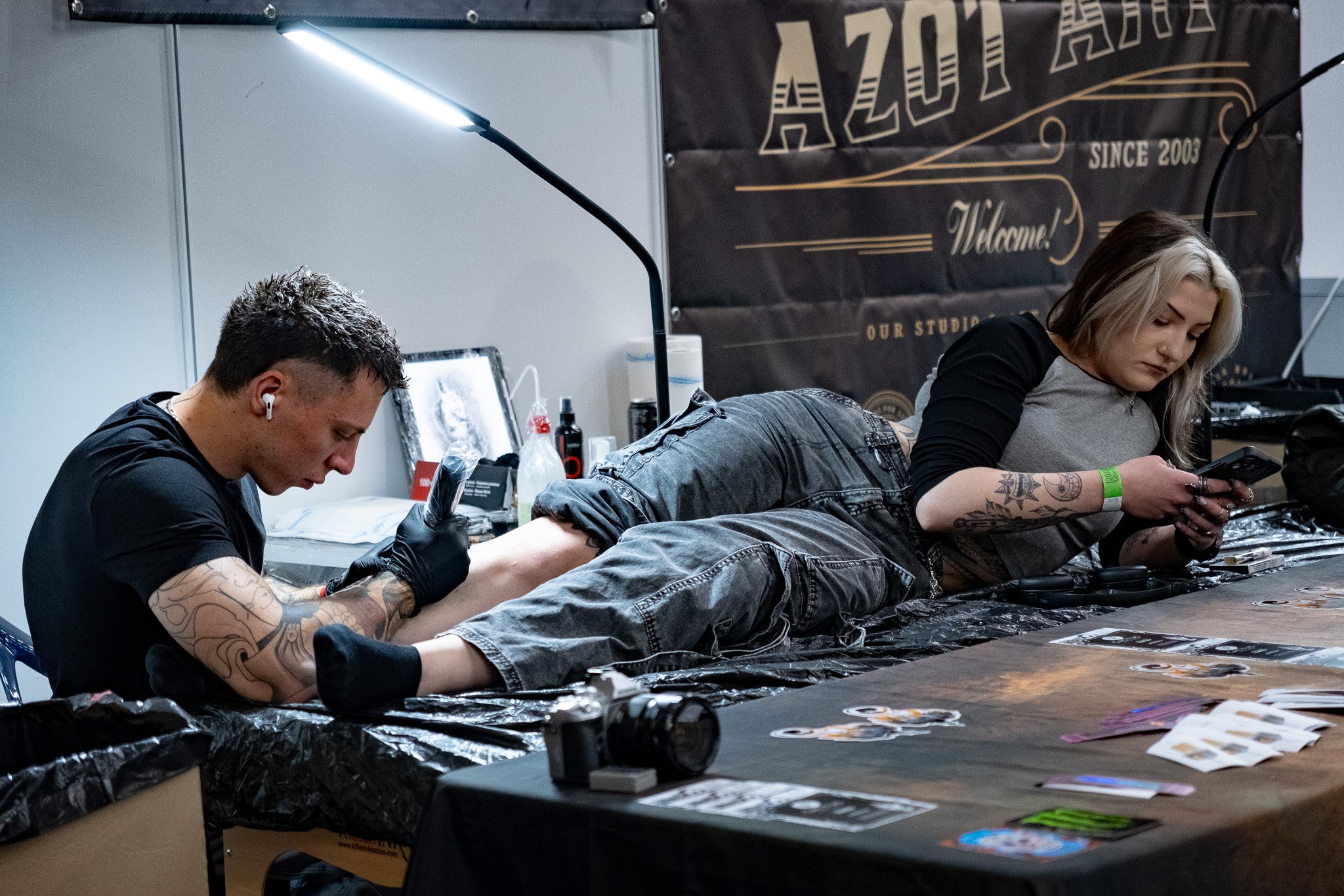 A person lying on a tattoo station table getting a tattoo on their leg while another person checks their phone. The tattoo artist is wearing gloves and wireless earbuds.